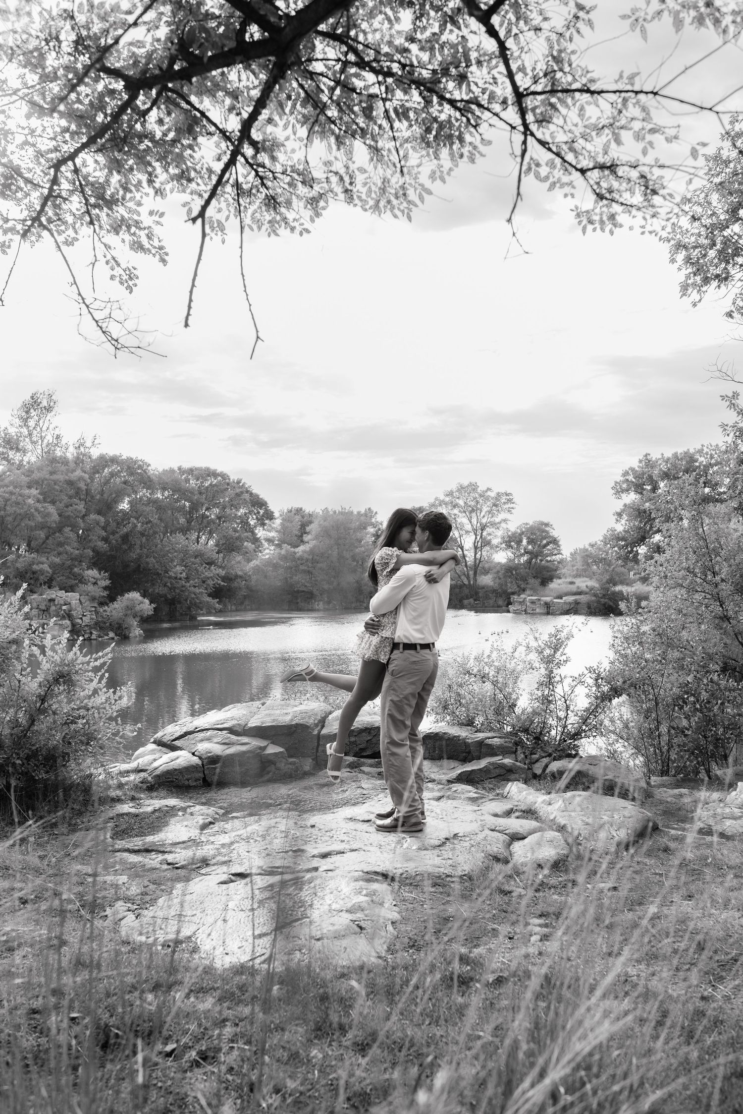 Black and white photo of a couple embracing romantically by a serene lake surrounded by lush trees.