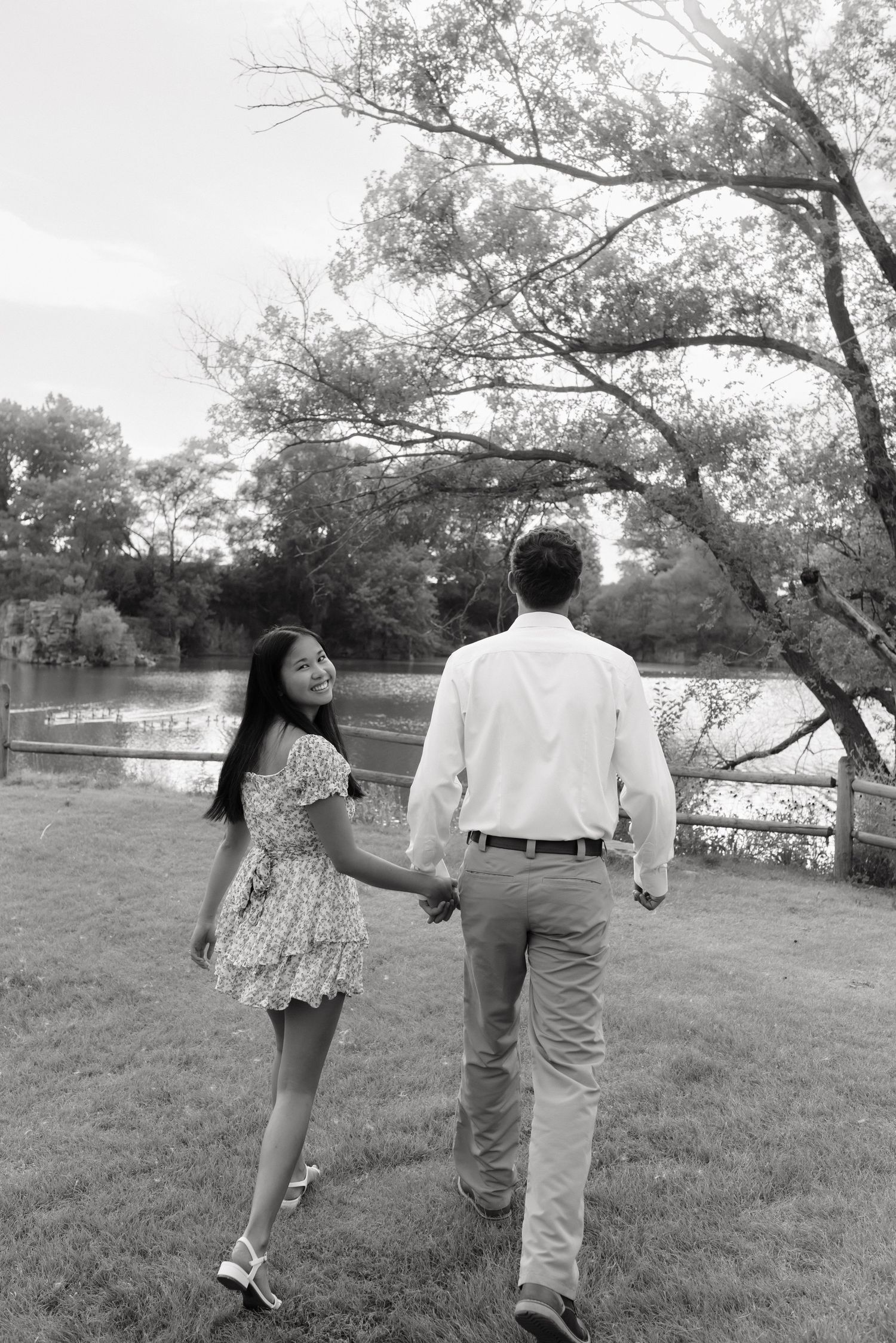 Black and white photo of a couple holding hands walking toward a serene lake surrounded by trees.