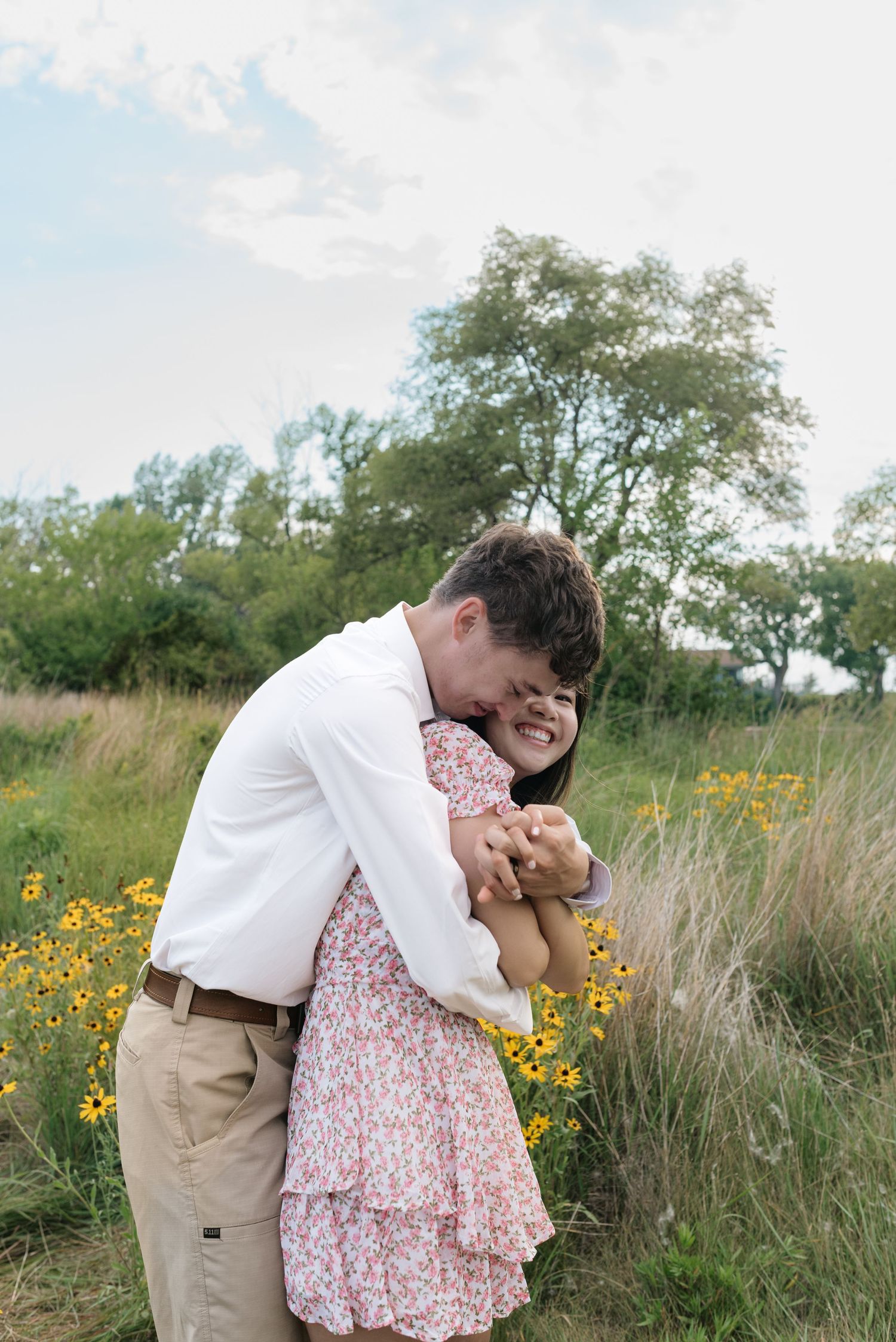 Young couple embracing and laughing in a wildflower field, man in white shirt hugging woman in floral dress.