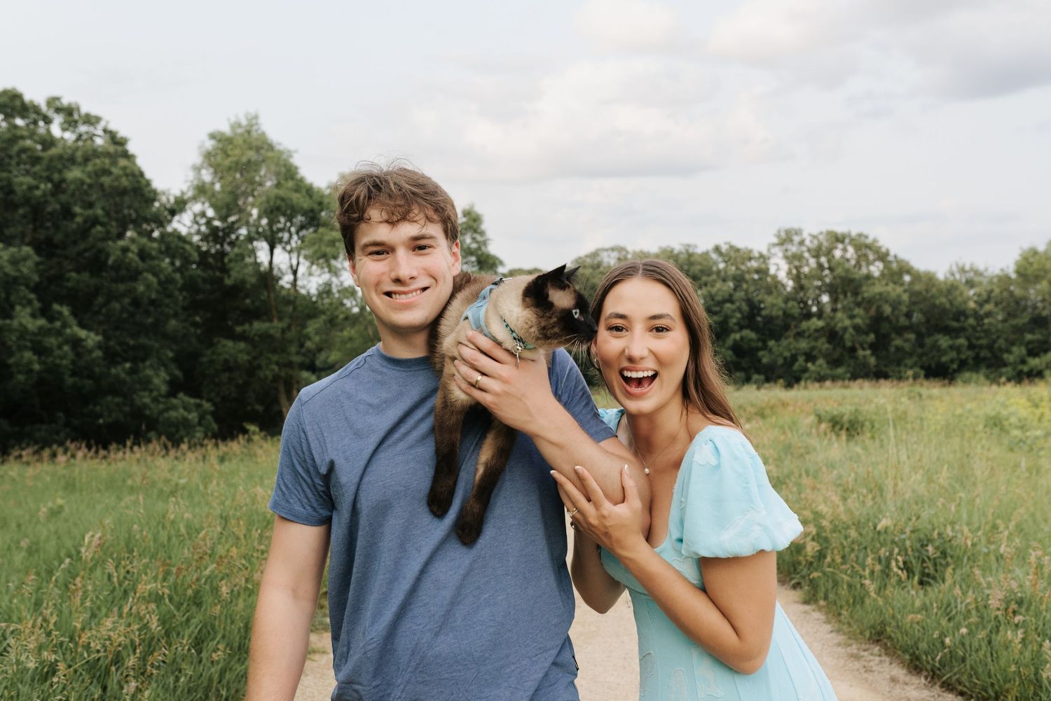 Young couple smiling outdoors holding a small baby goat on a scenic dirt path surrounded by green fields.