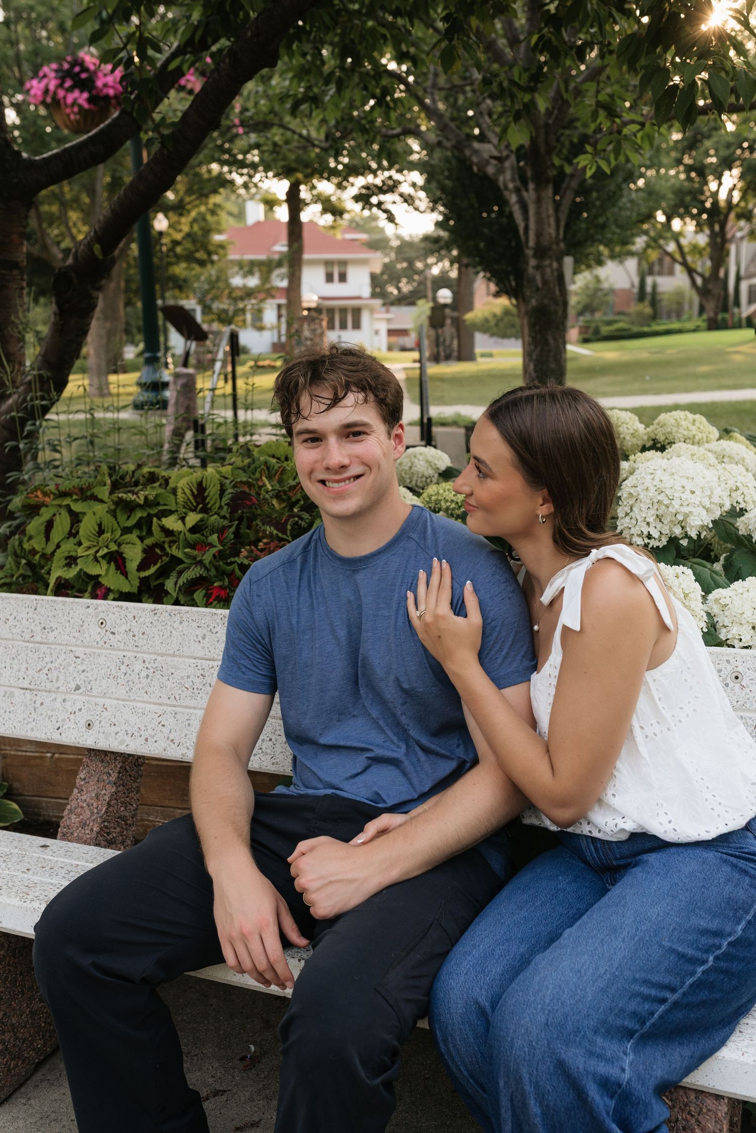 A smiling couple sits on a stone bench in a lush park, surrounded by flowers and greenery.