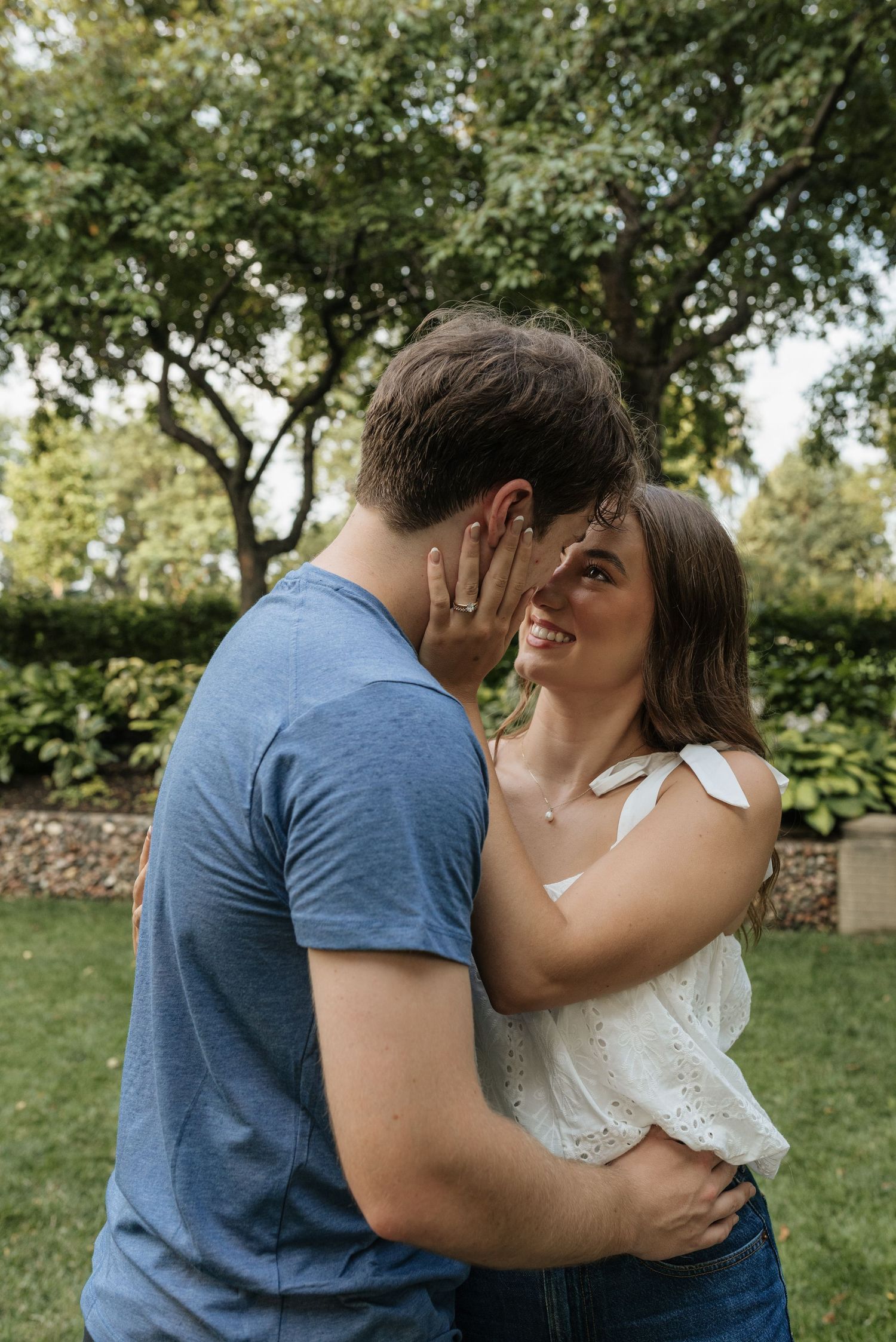 Happy couple embracing and almost kissing in a garden, woman laughing joyfully, wearing white top and necklace.