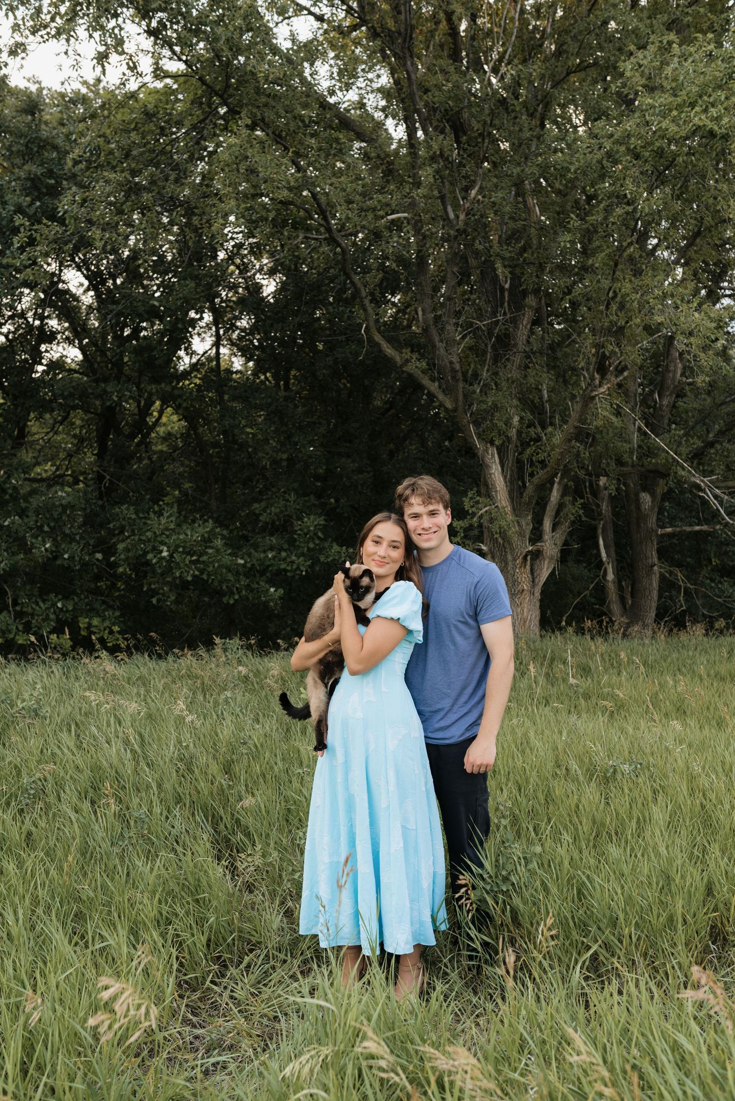 A couple stands in a lush green field holding a black cat, woman in light blue dress, man in blue t-shirt.
