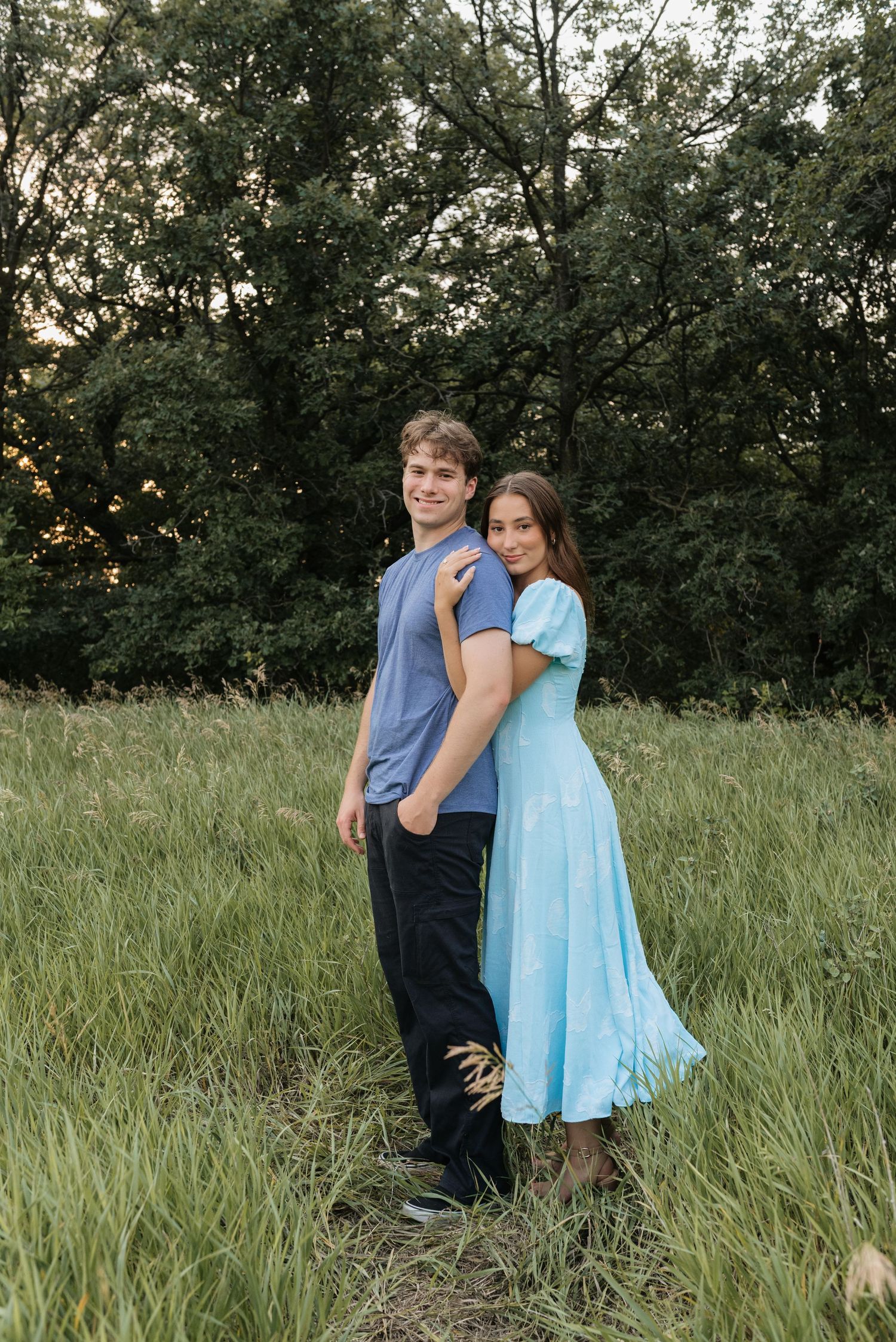 A young couple poses in a lush green field, woman in light blue dress embracing man in blue shirt and black pants.