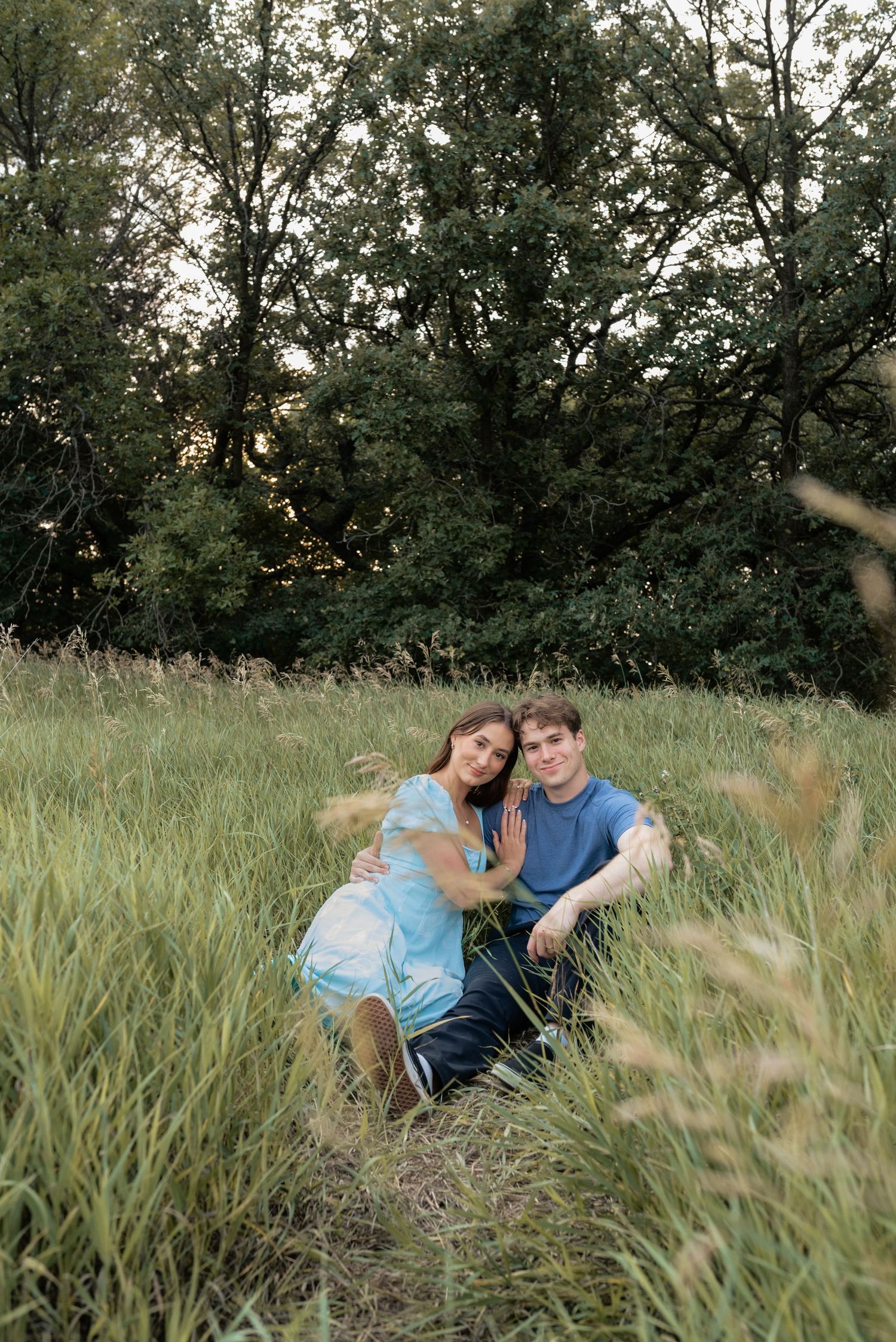 A couple sits together in a lush green meadow, smiling warmly, dressed in matching blue outfits with trees behind.