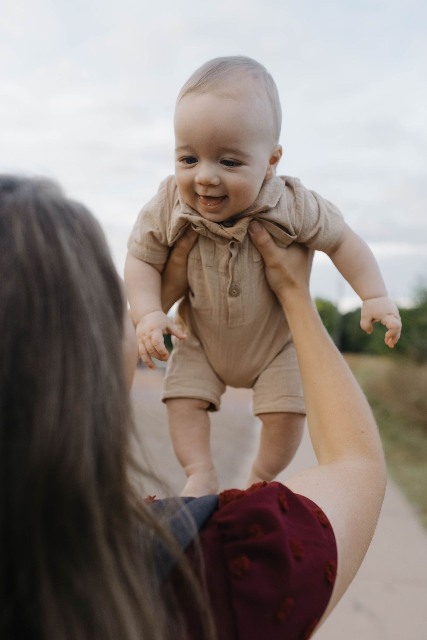 Happy baby in tan romper being held up by parent outdoors, smiling on a peaceful nature path.