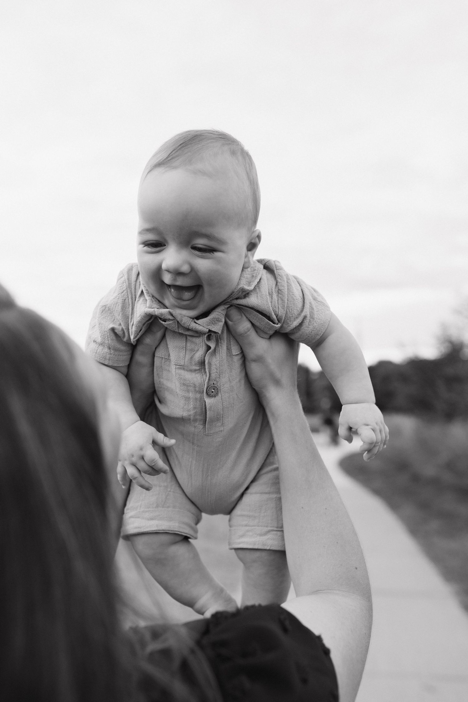Black and white photo of a giggling baby being lifted by a woman outside on a bright day.