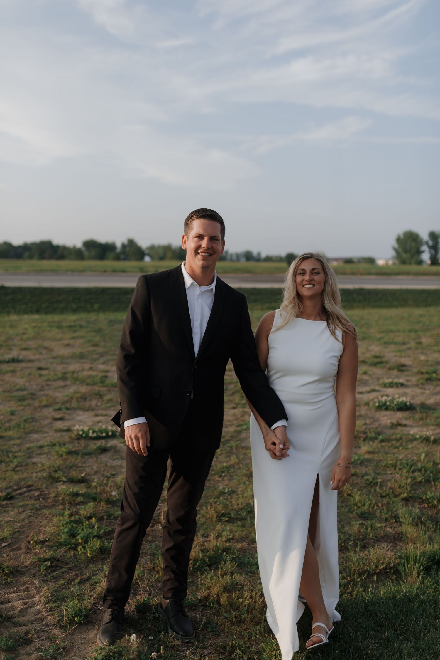 Happy couple posing together in wedding attire on a green field with a bright sky backdrop.