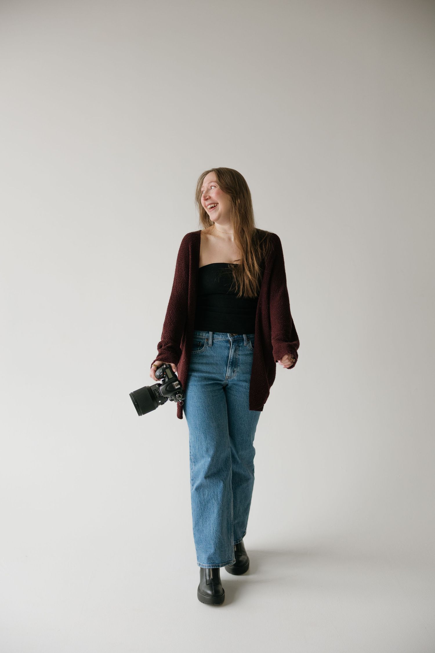 Smiling female photographer walking with camera in hand wearing burgundy cardigan and blue jeans in studio.