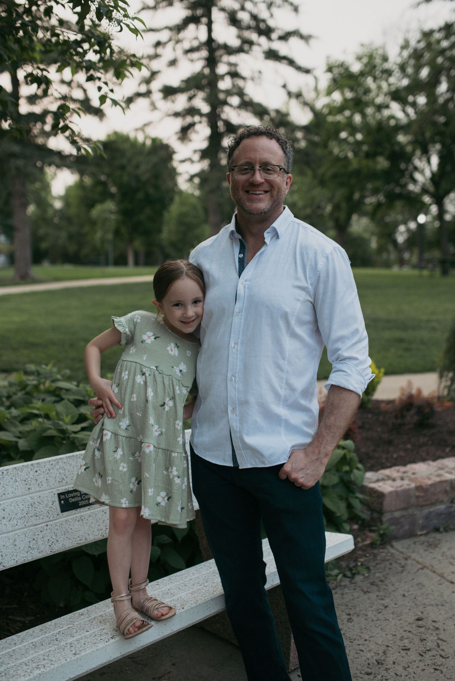 Smiling father and daughter in green floral dress posing together on a park bench.