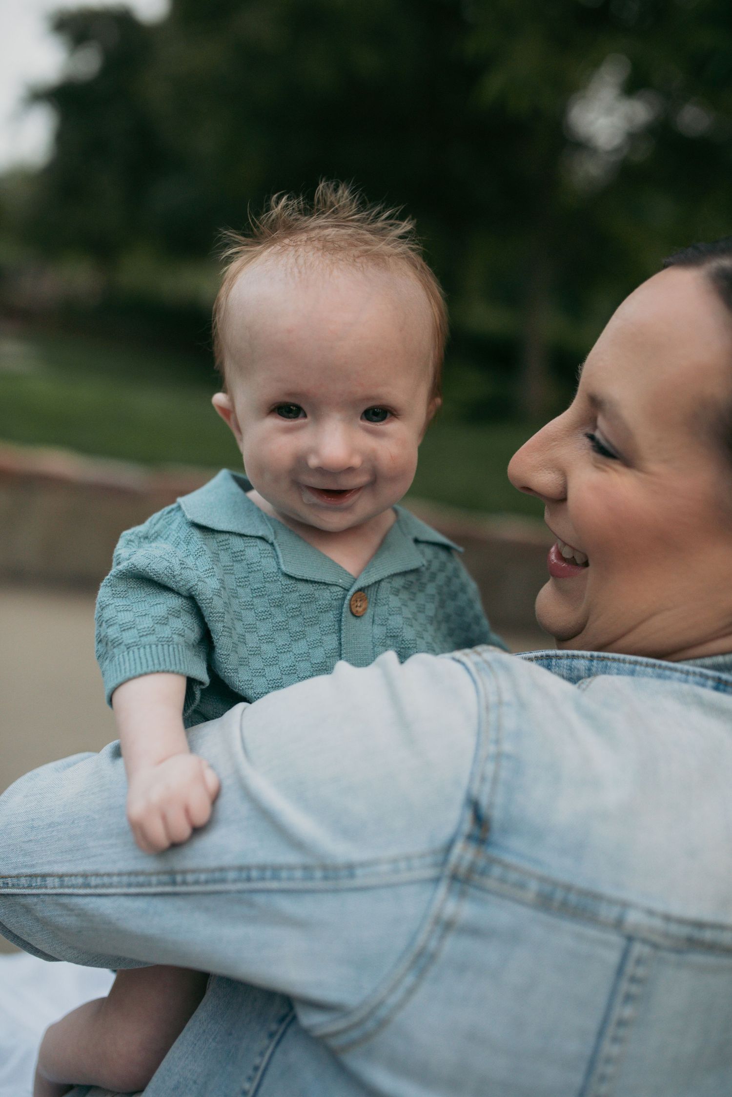 A happy baby in a teal checkered knit romper smiling while being held by a woman outdoors.