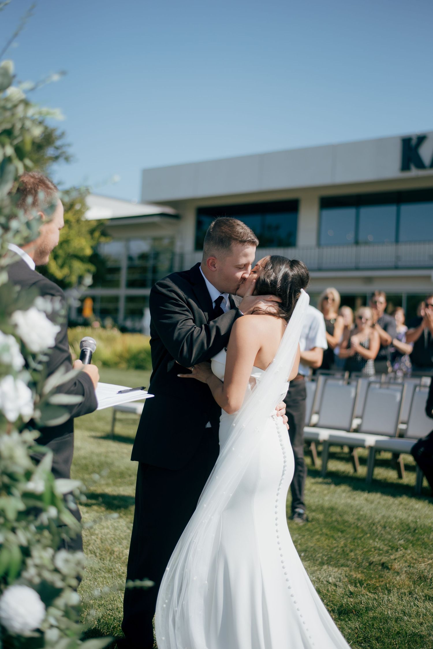 Newlyweds share first kiss at outdoor wedding ceremony while guests applaud in sunny celebration.