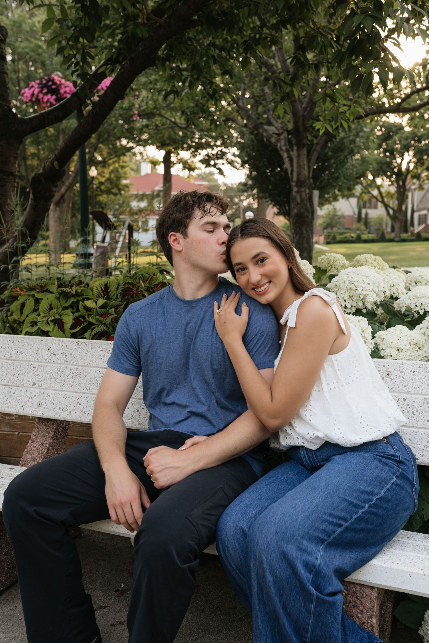A couple sits on a white bench in a garden, man kissing woman's temple as she smiles, surrounded by lush greenery.