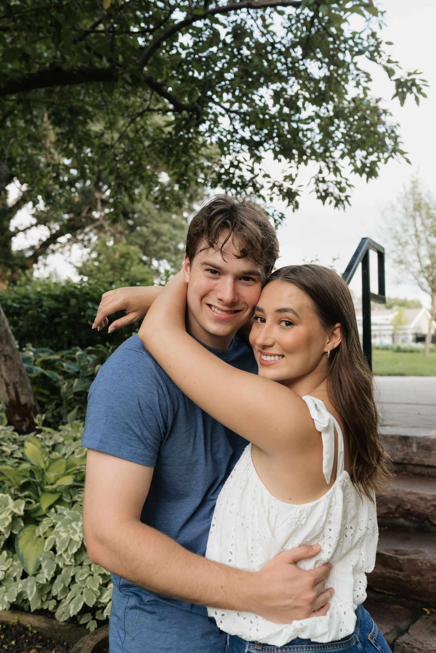 A smiling young couple embracing outdoors, with lush green trees and garden in the background.