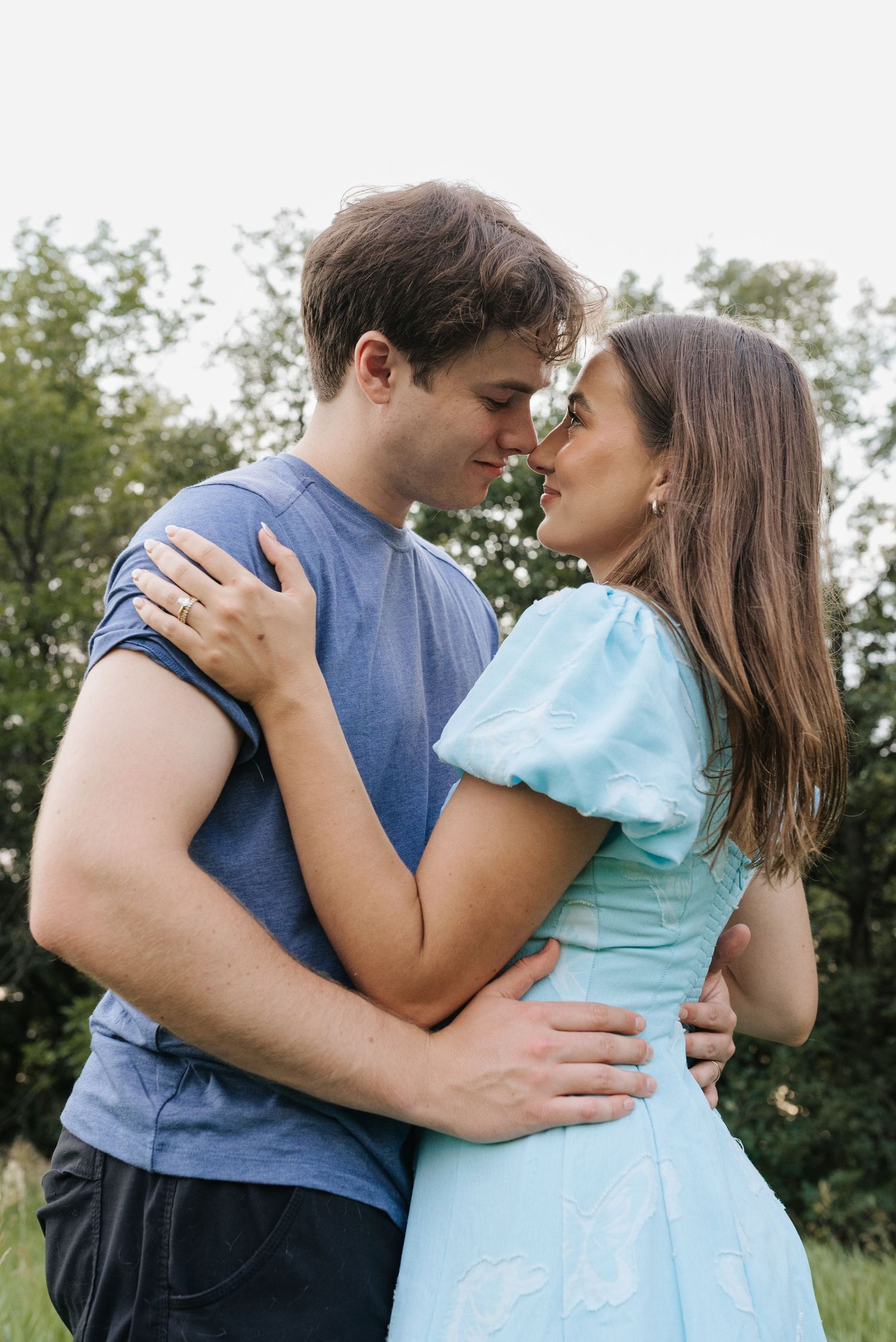 A young couple embraces outdoors, nearly kissing, with the woman in a light blue dress and man in a blue t-shirt.
