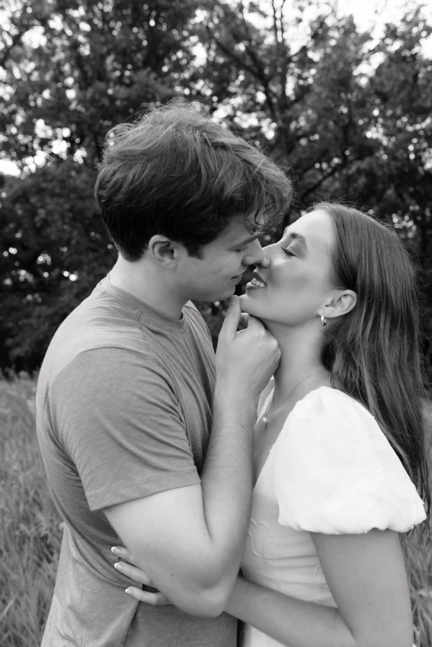 Black and white photo of a romantic couple almost kissing outdoors, surrounded by lush trees in a natural setting.