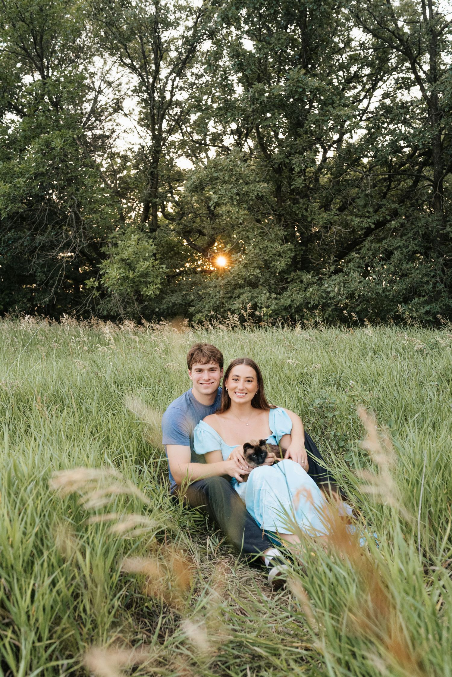 A couple sits together in a lush green meadow at golden hour, smiling warmly while holding a small dog.
