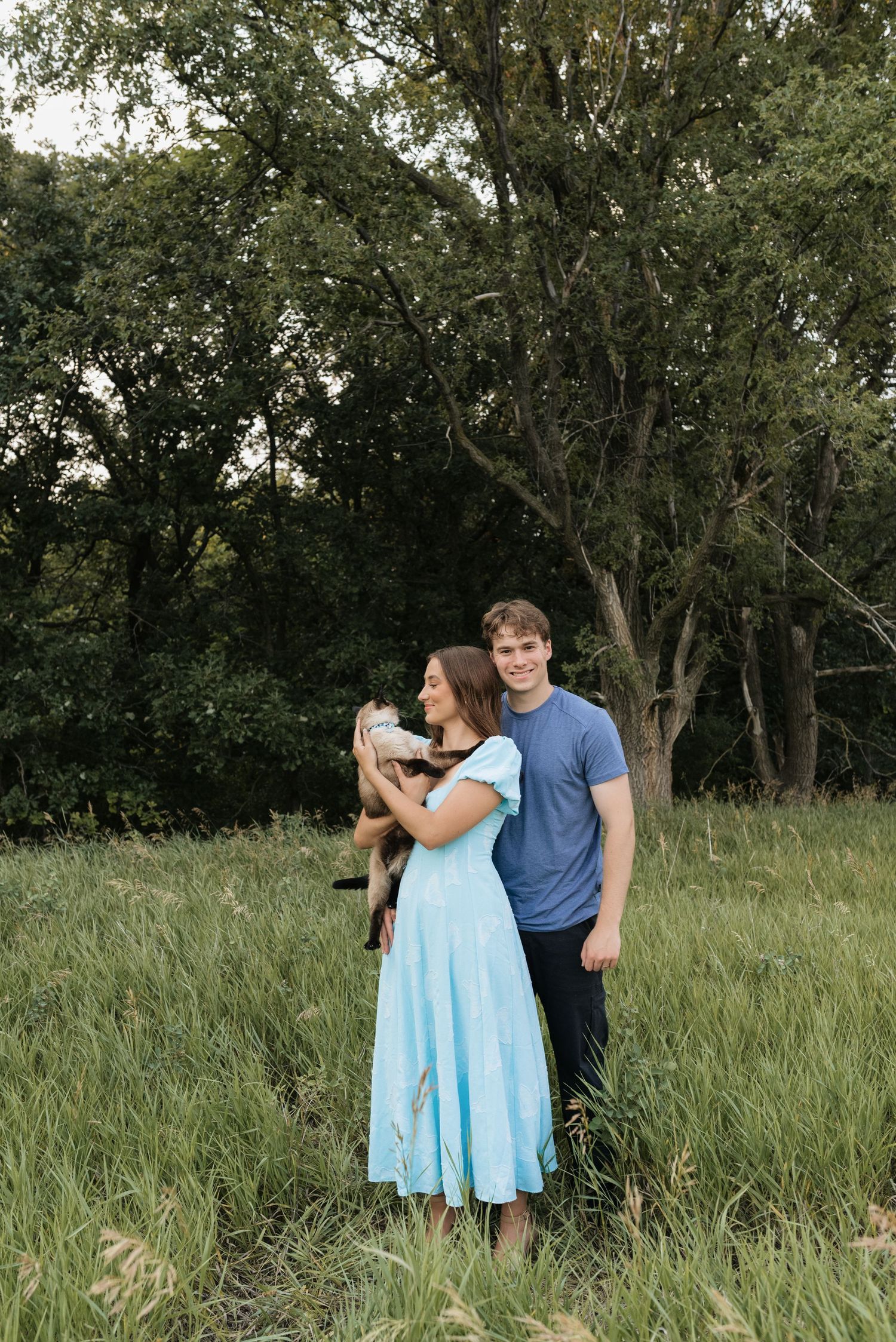 A couple stands in a lush green field, woman in light blue dress holding a puppy, man in blue t-shirt smiling.