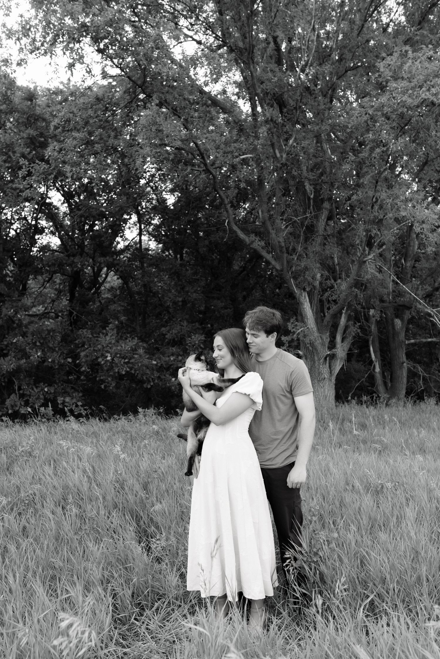 Black and white photo of a couple standing in a grassy field holding a small dog, with trees in the background.