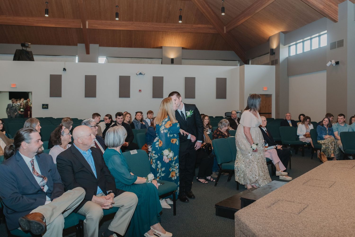 Wedding guests seated in a church ceremony hall as the groom escorts a guest to her seat.