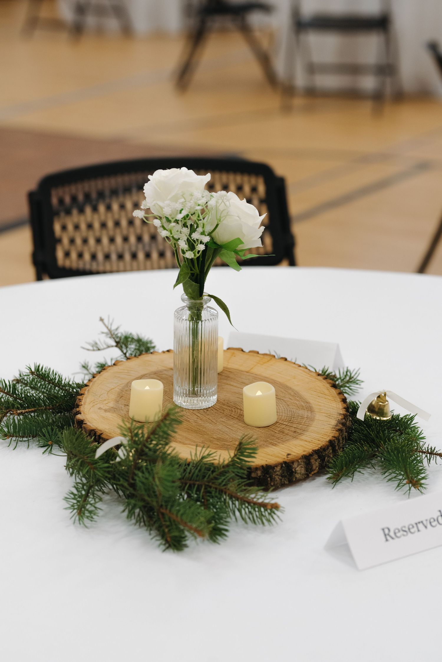 Rustic wedding centerpiece with white flowers in glass vase on wood slice with greenery and candles.