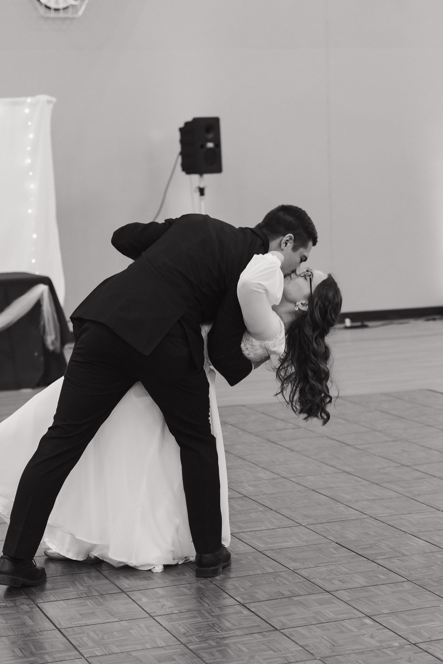 Black and white photo of bride and groom sharing romantic dip kiss during first dance at wedding reception.