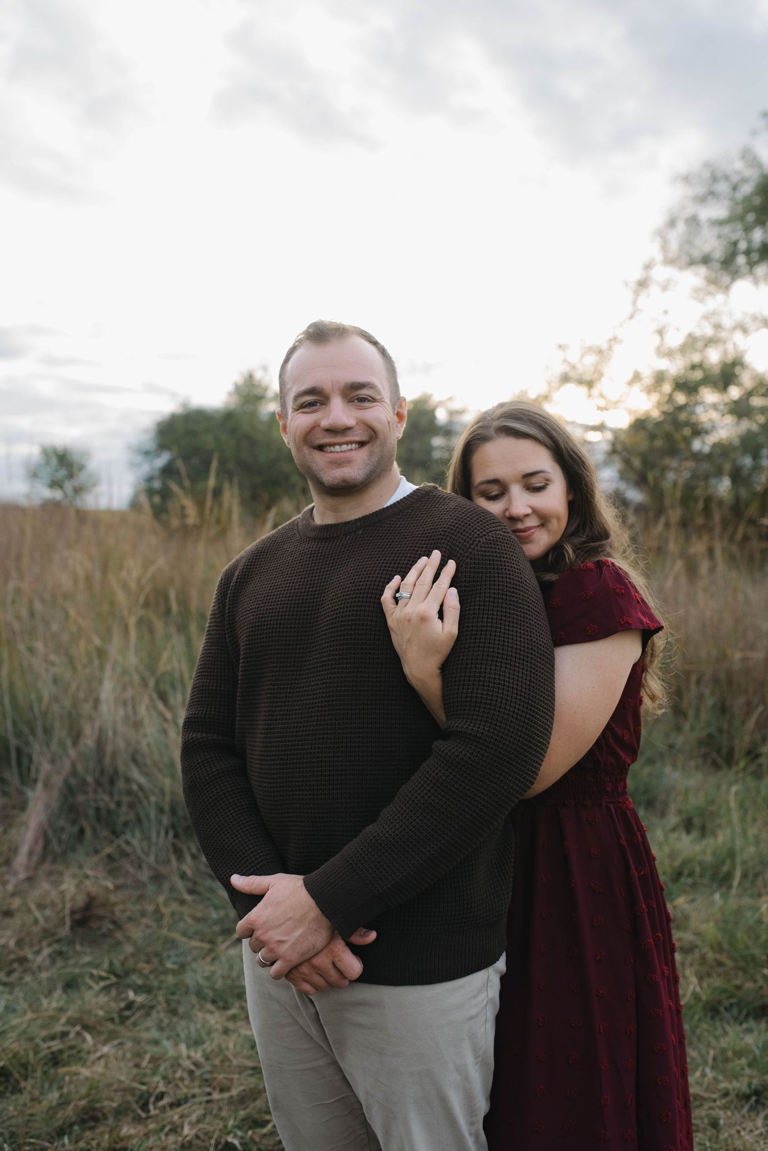 A woman in burgundy embraces a smiling man from behind in a golden grass field at sunset.