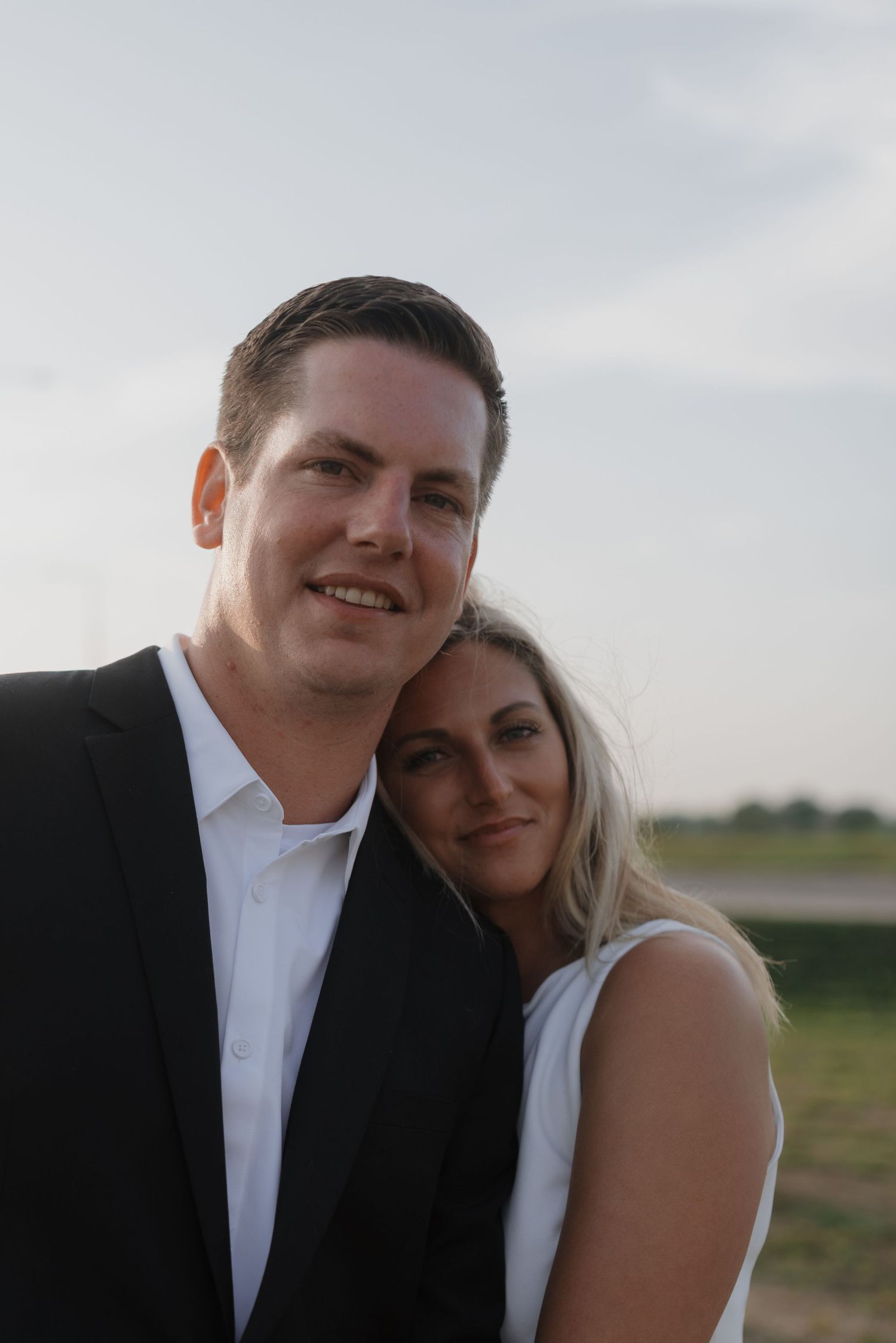 Smiling couple poses outdoors at dusk, woman in white dress leaning on man in black suit.