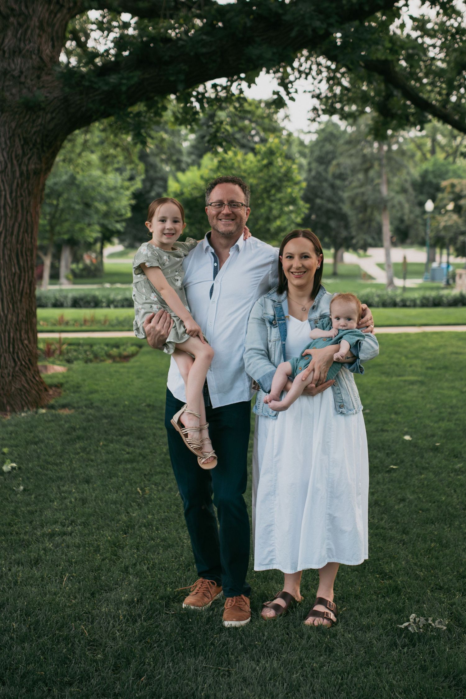 Family portrait of four under a large tree in a park, parents holding two young children.