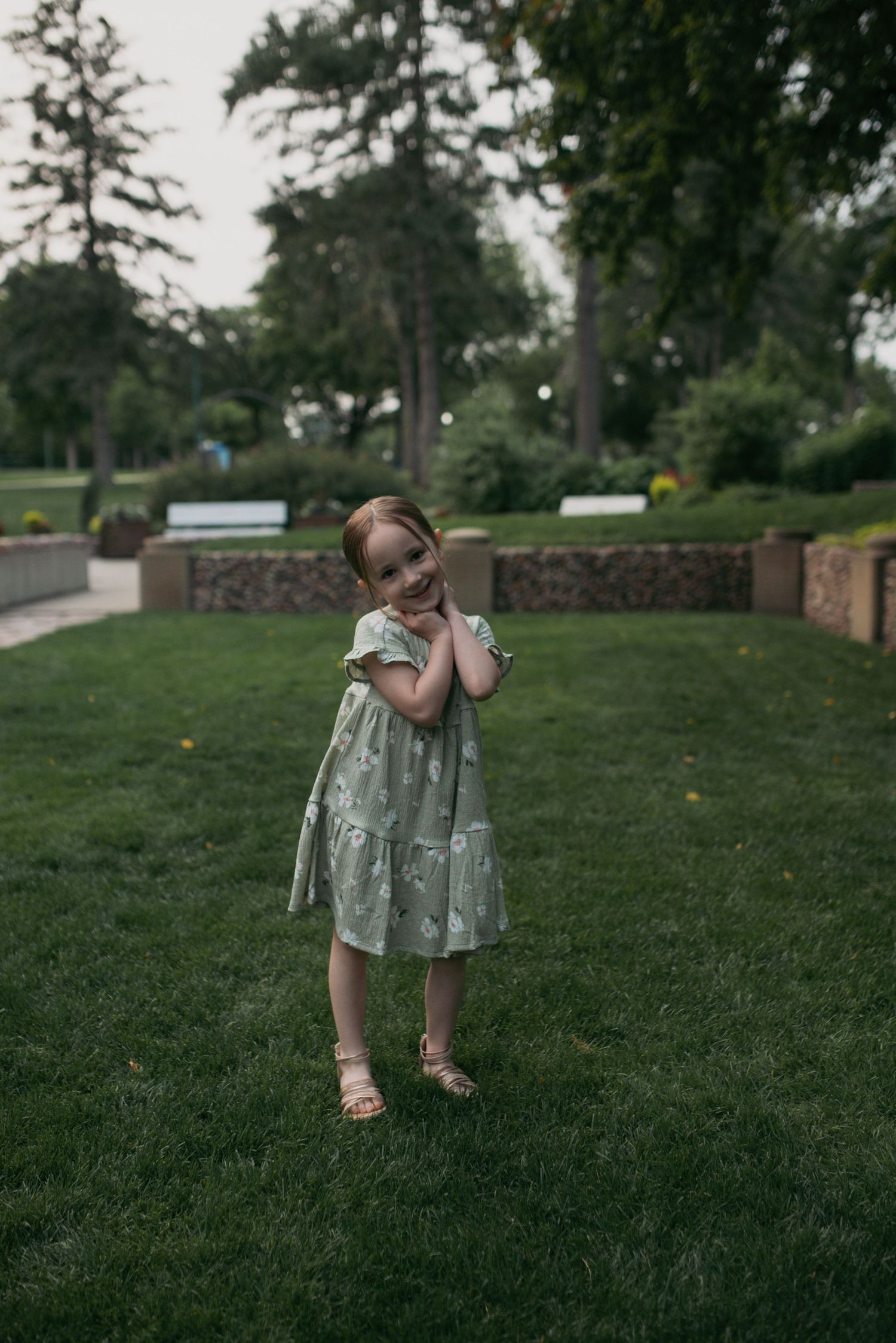 Adorable young girl in sage green floral dress smiling shyly in a lush park.