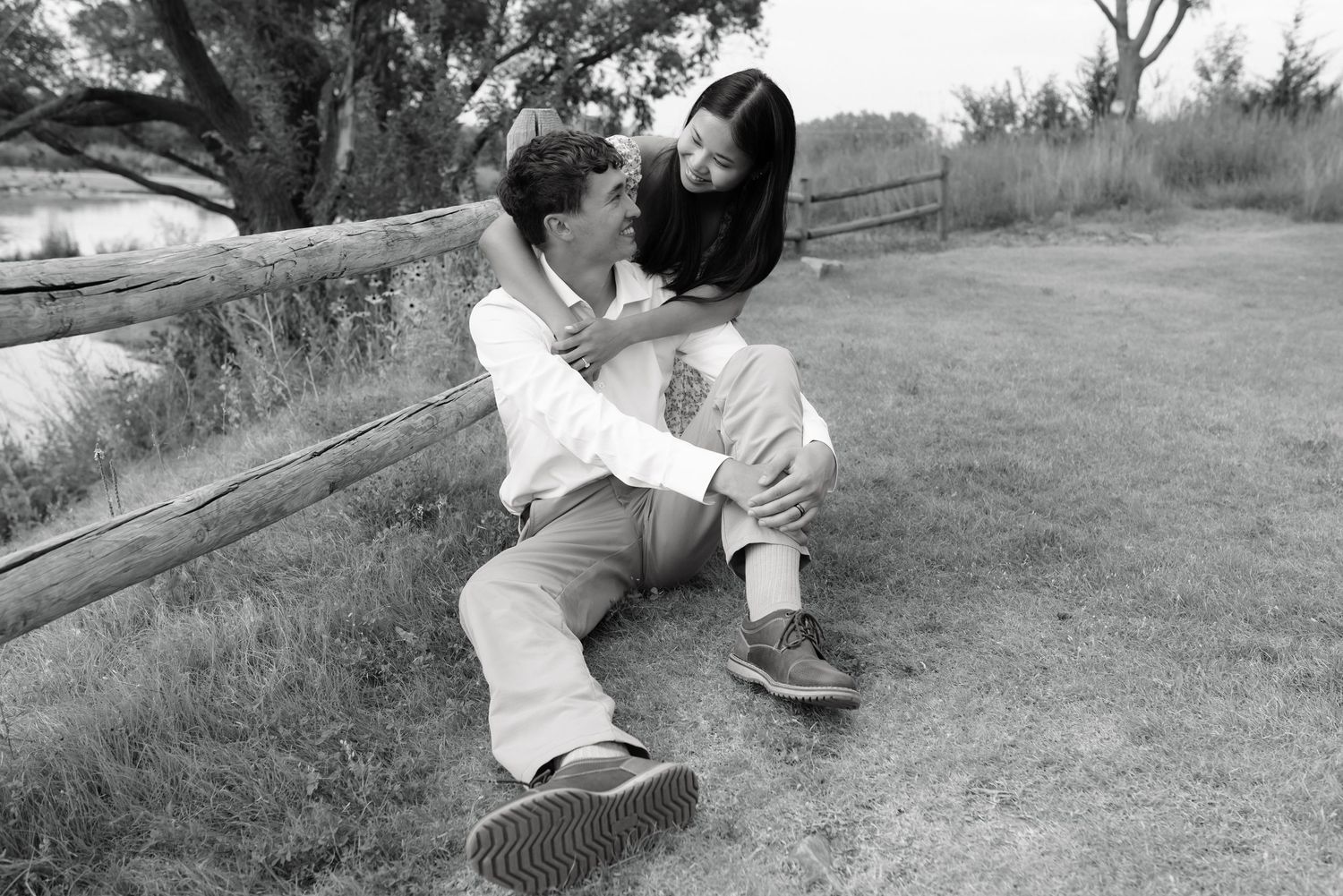 Black and white photo of a couple sitting together on grass near a wooden fence in a romantic outdoor setting.