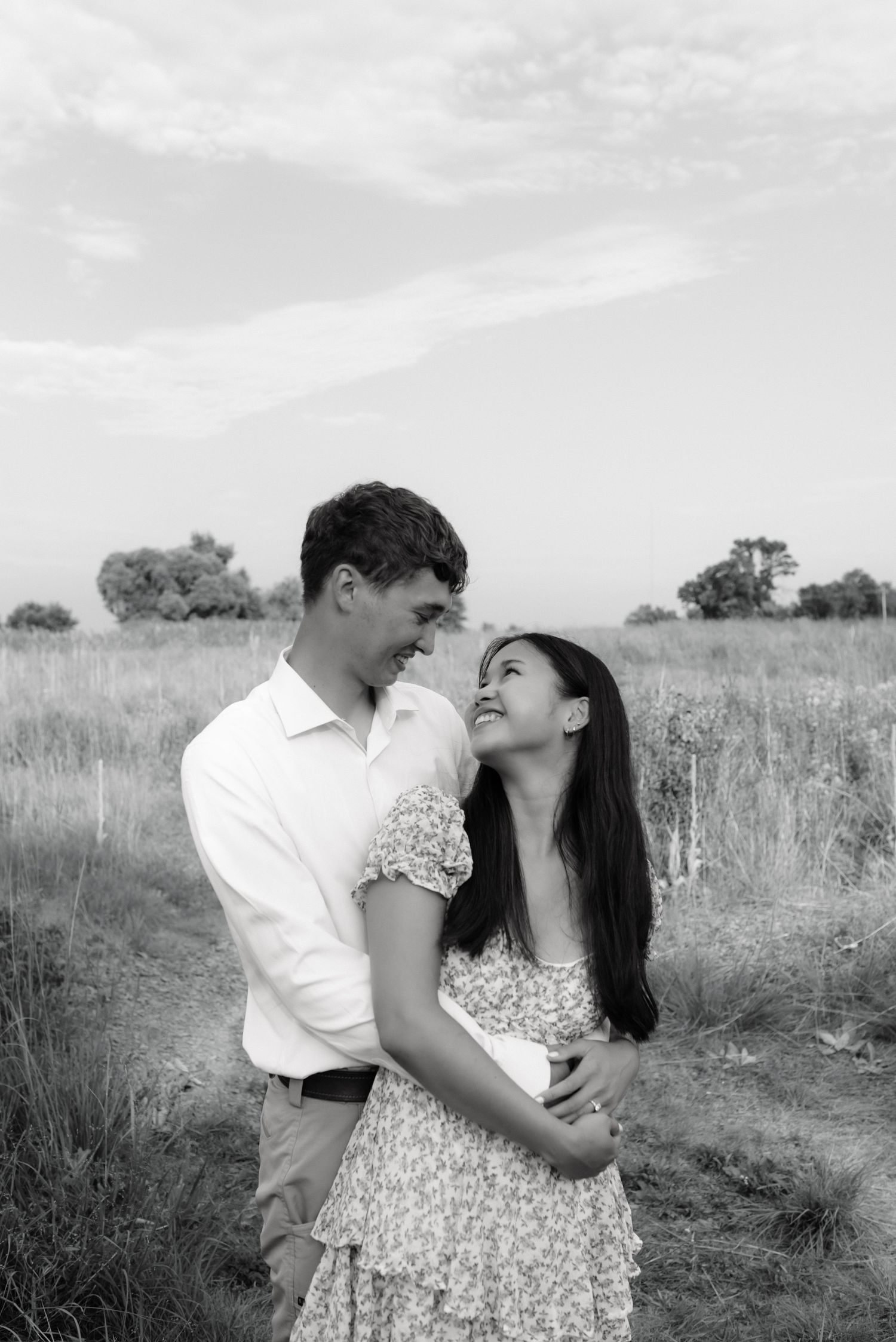 Black and white photo of smiling couple embracing on a rural path with open fields.