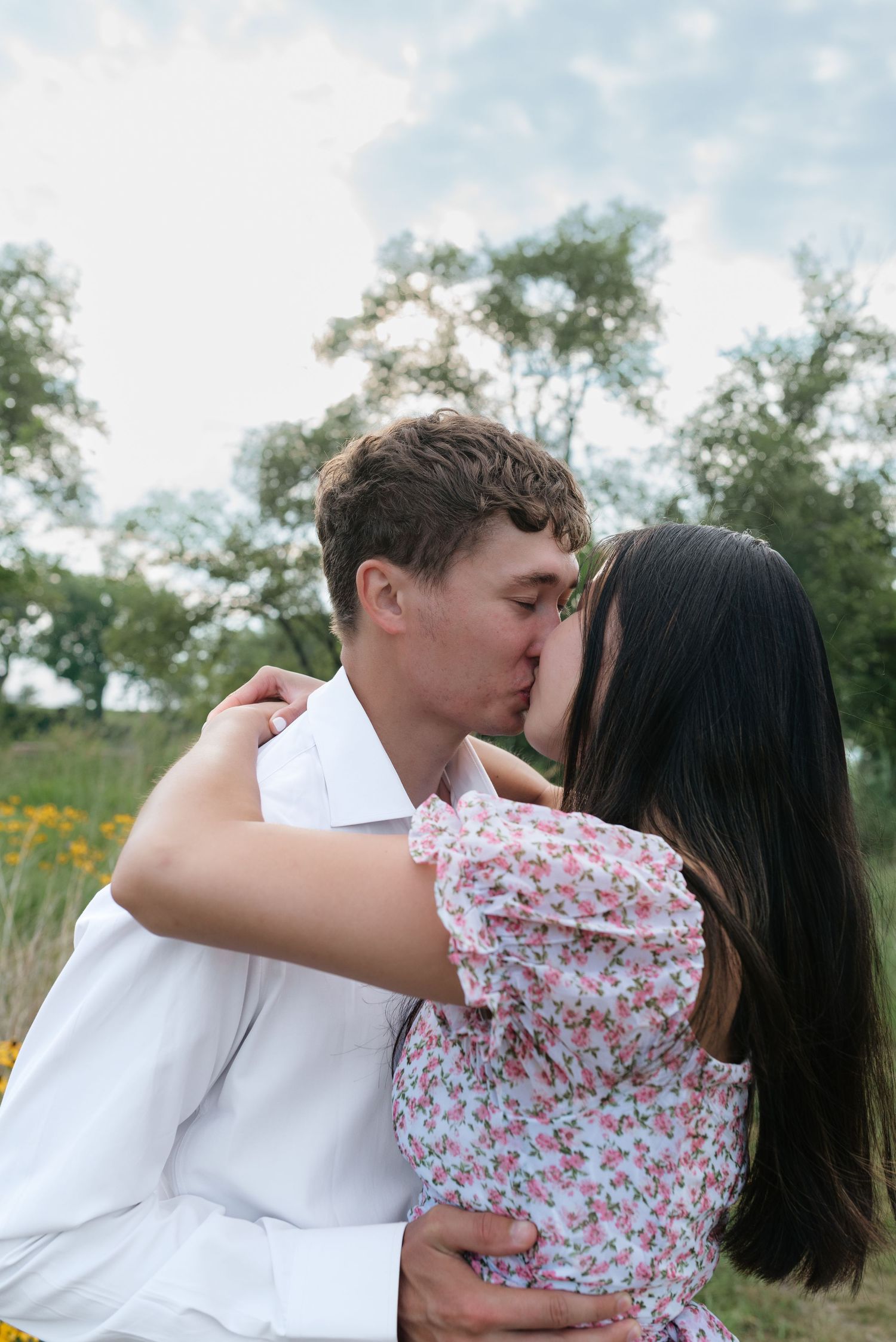 Young couple sharing a romantic kiss outdoors in a green field under a cloudy sky.