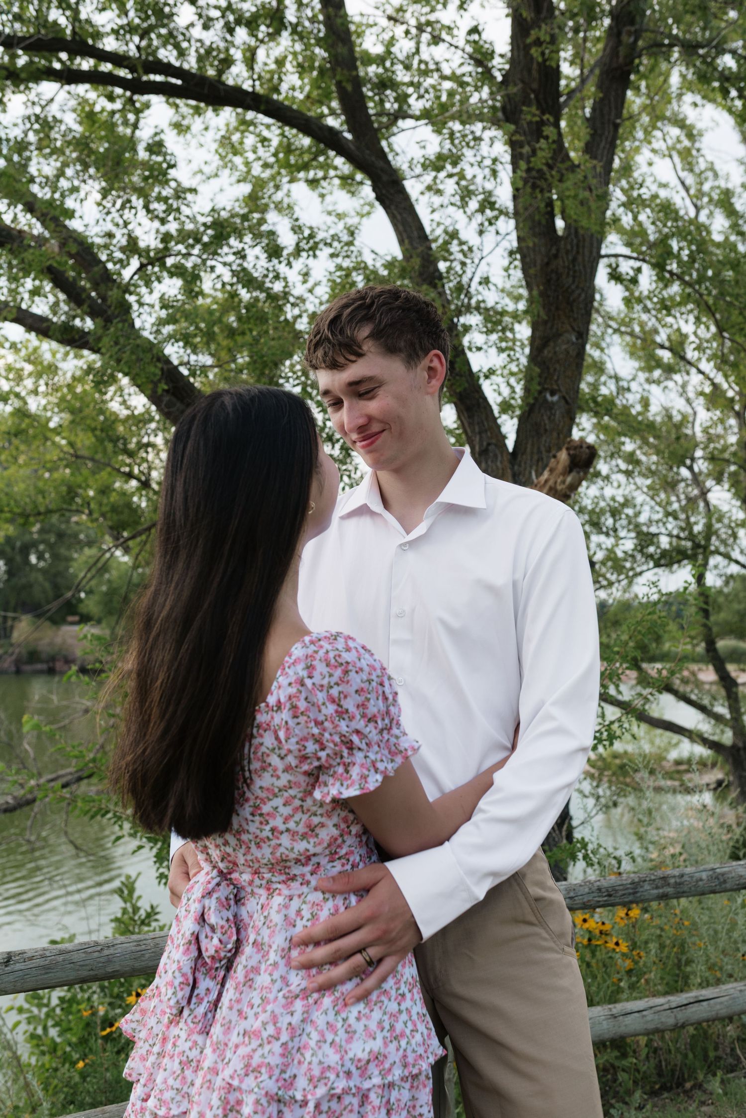 Young couple embracing and smiling at each other near a wooden fence with a tranquil pond backdrop.