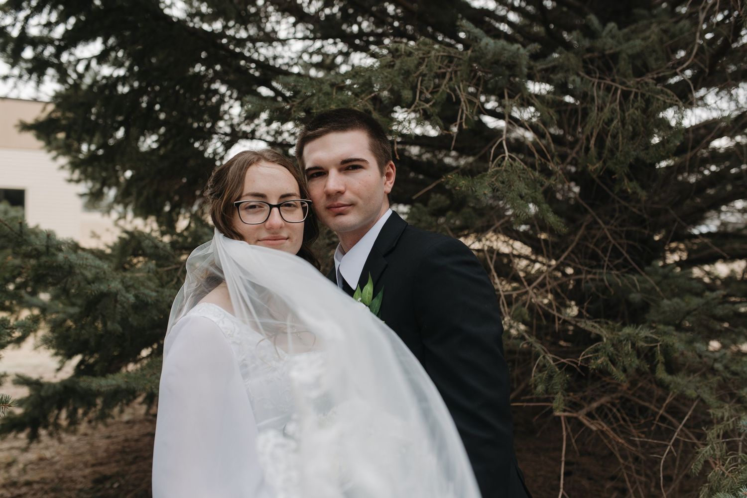 Bride in white veil and glasses embraces groom in dark suit outdoors near evergreen trees.