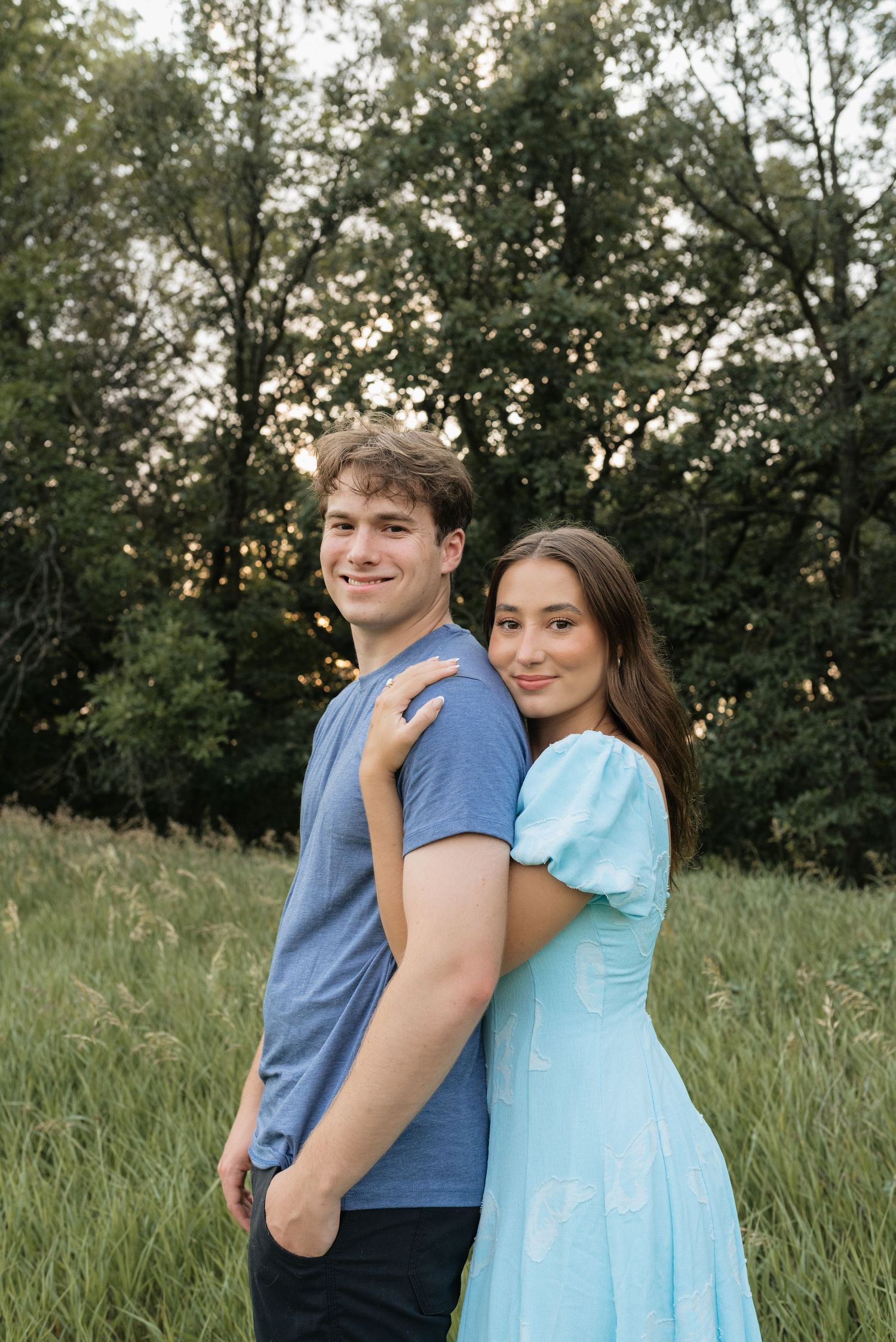 A young couple poses in a green field, the woman in a light blue dress resting her hand on the man's shoulder.