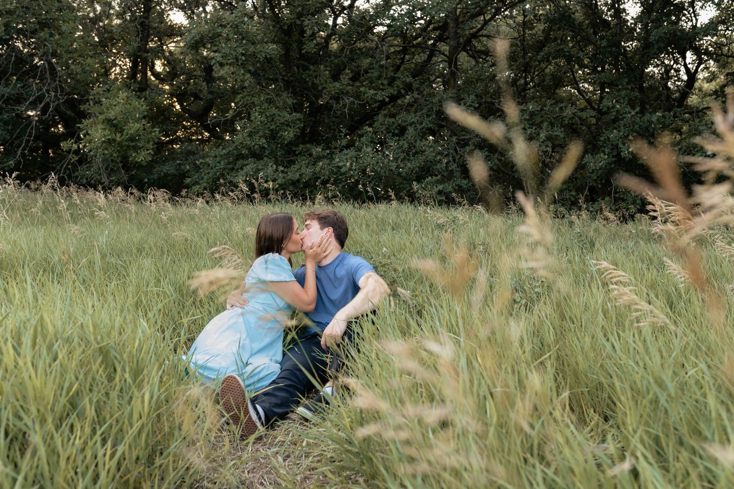 Couple sharing a romantic kiss while sitting in a lush green meadow surrounded by tall grass and trees.