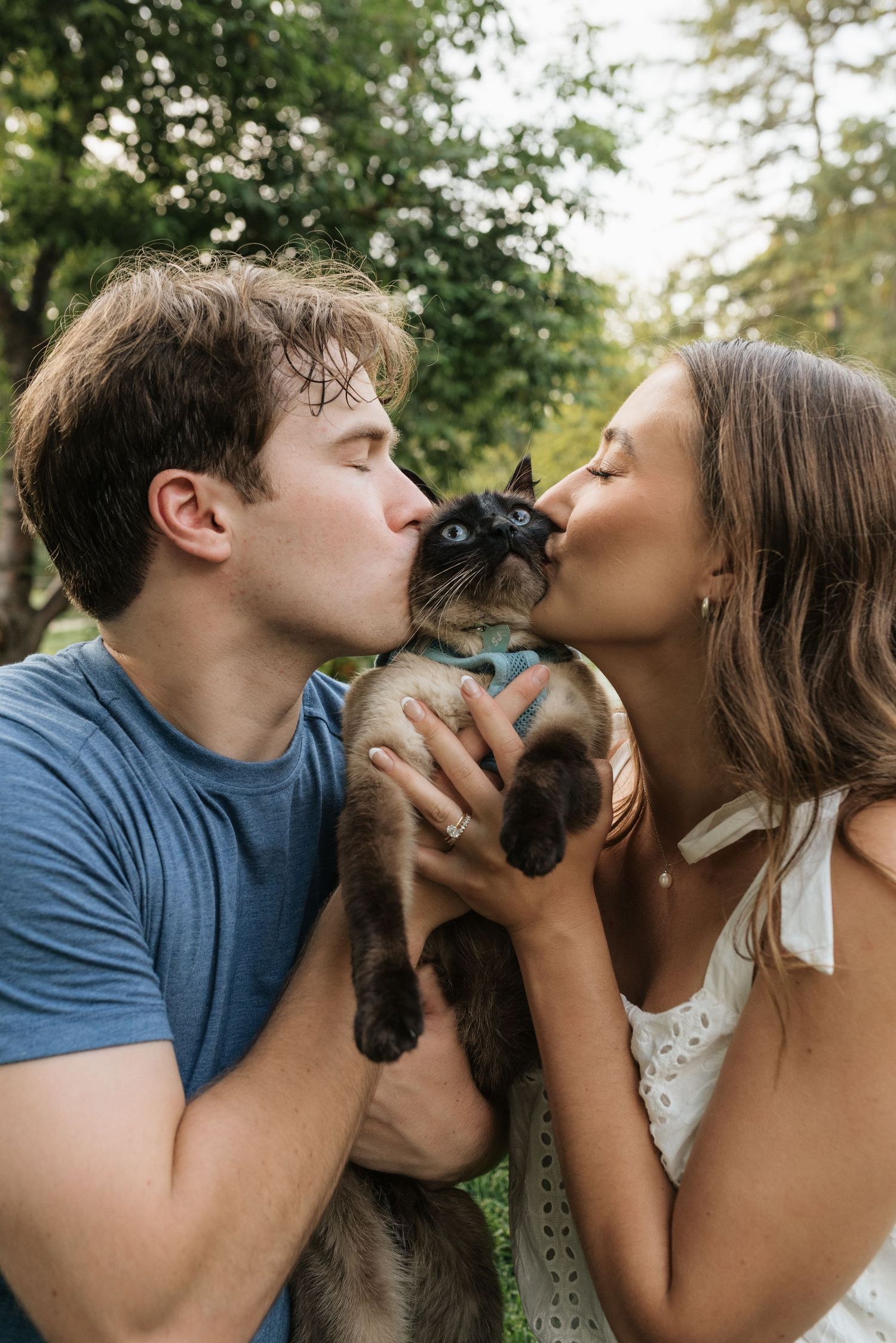 A couple kisses their Siamese cat outdoors, surrounded by lush green trees in a heartwarming pet portrait.