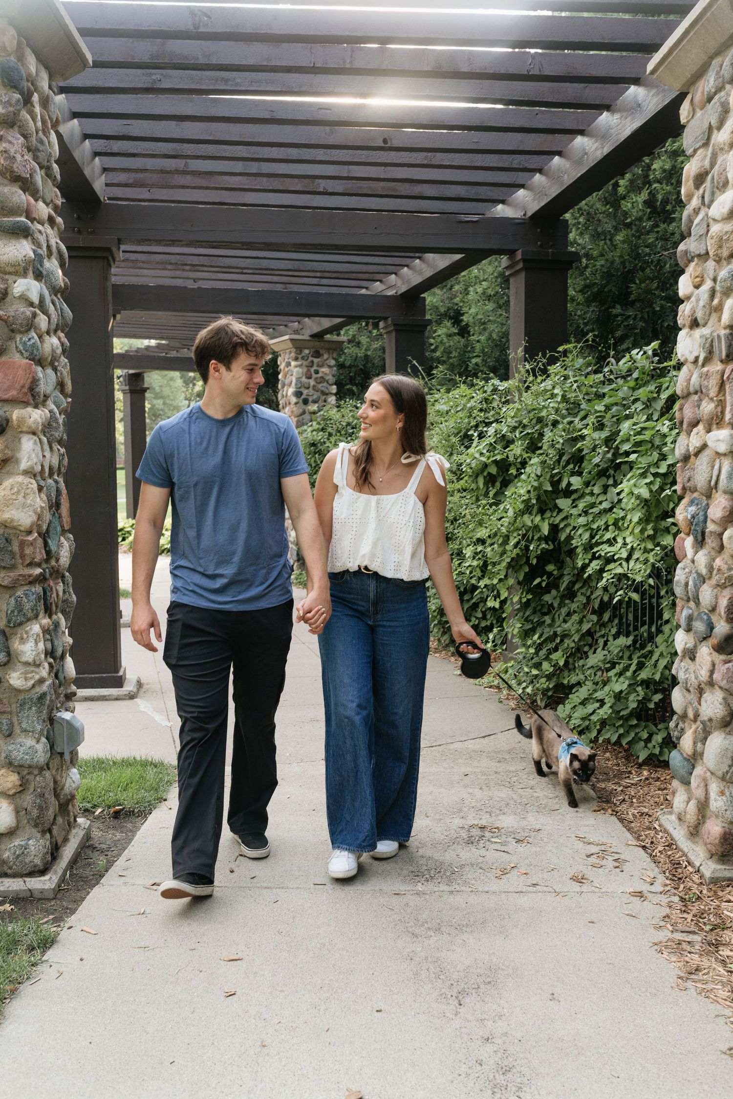 Young couple smiling at each other while walking their small dog under a wooden pergola with stone columns.