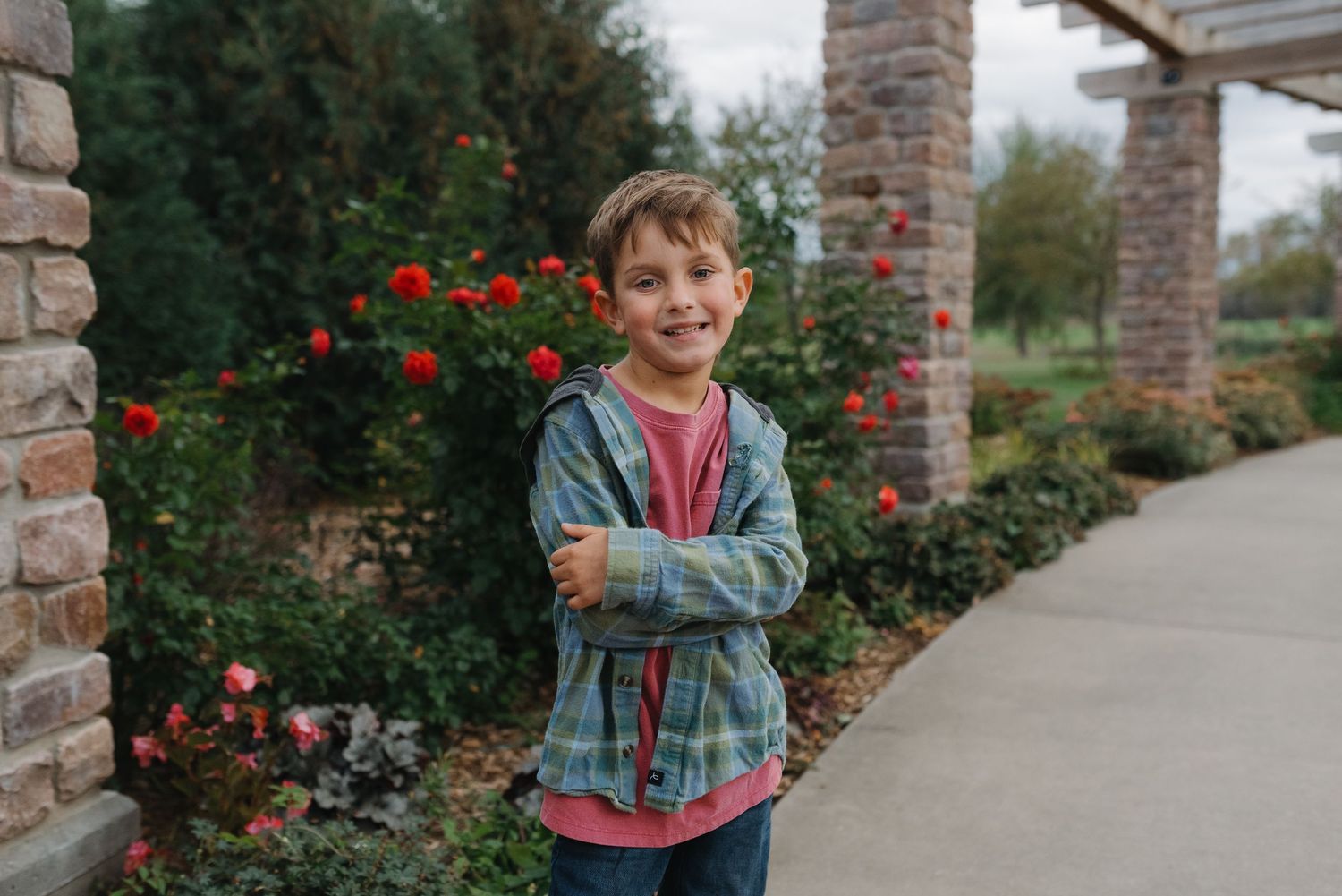 Smiling boy in a plaid hoodie standing with arms crossed in a garden with red roses.