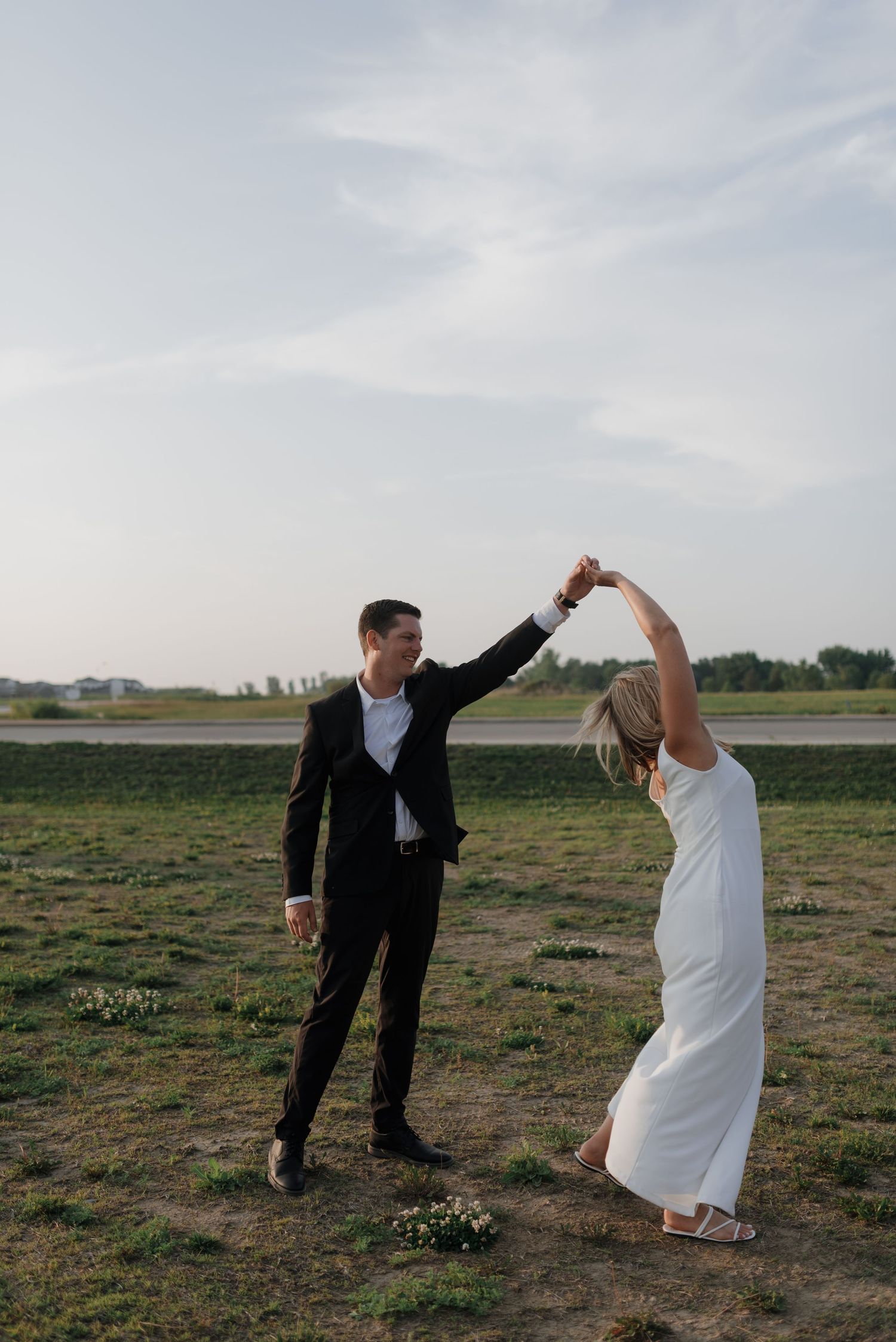 Man in black suit spinning woman in white dress while dancing outdoors in open green field.