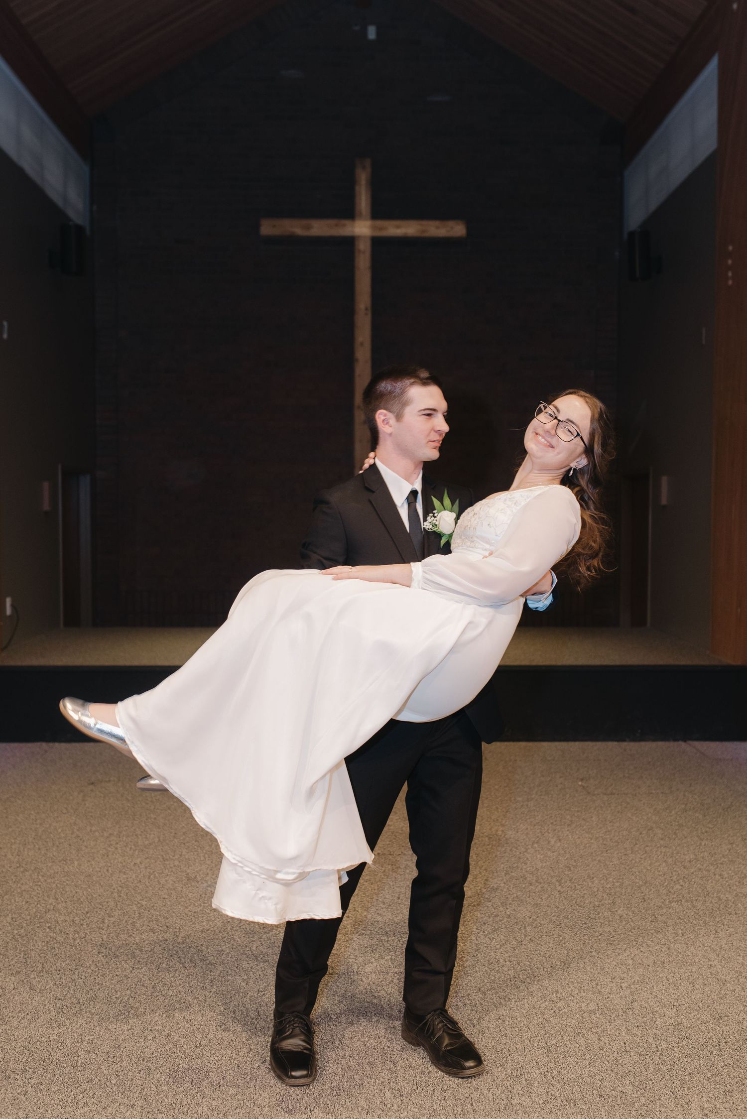 Groom carrying bride in white wedding dress inside a church with a large wooden cross in the background.