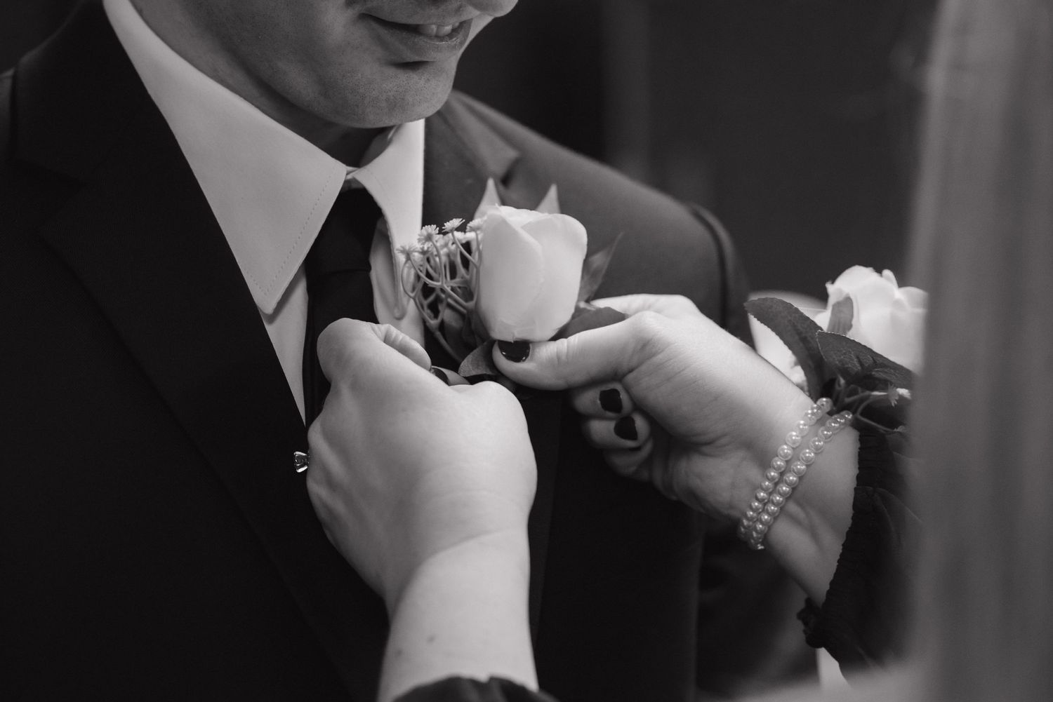 Black and white photo of a boutonniere being pinned onto a grooms suit lapel at a wedding.