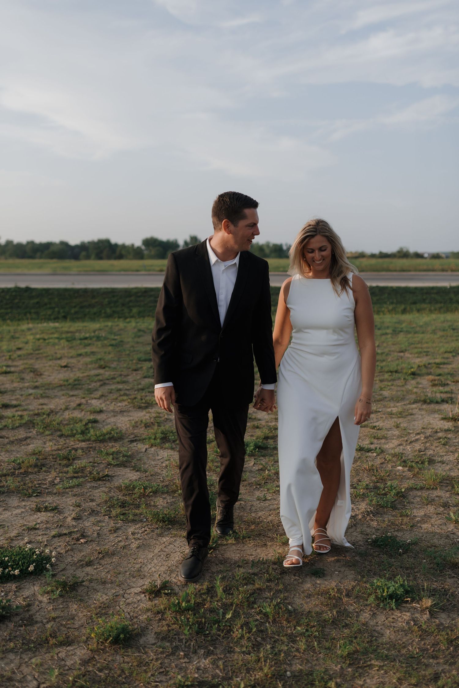 Smiling bride in a white slit gown and groom in black suit walking hand in hand outdoors.