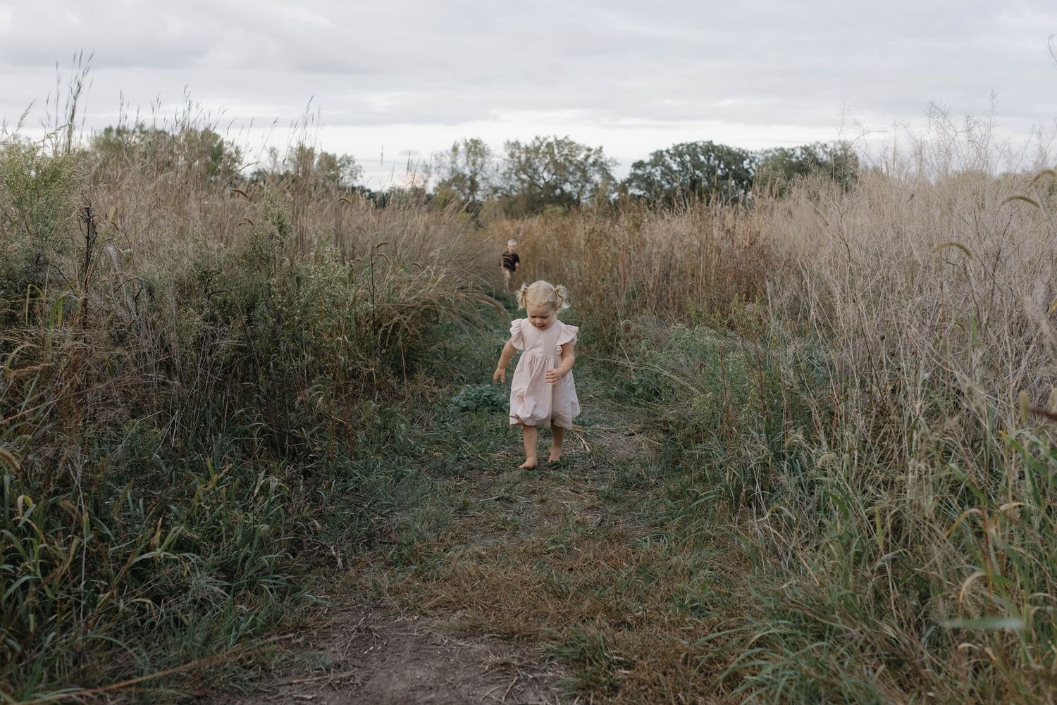 A toddler girl in a light pink dress stands on a grassy trail surrounded by tall wild grass.
