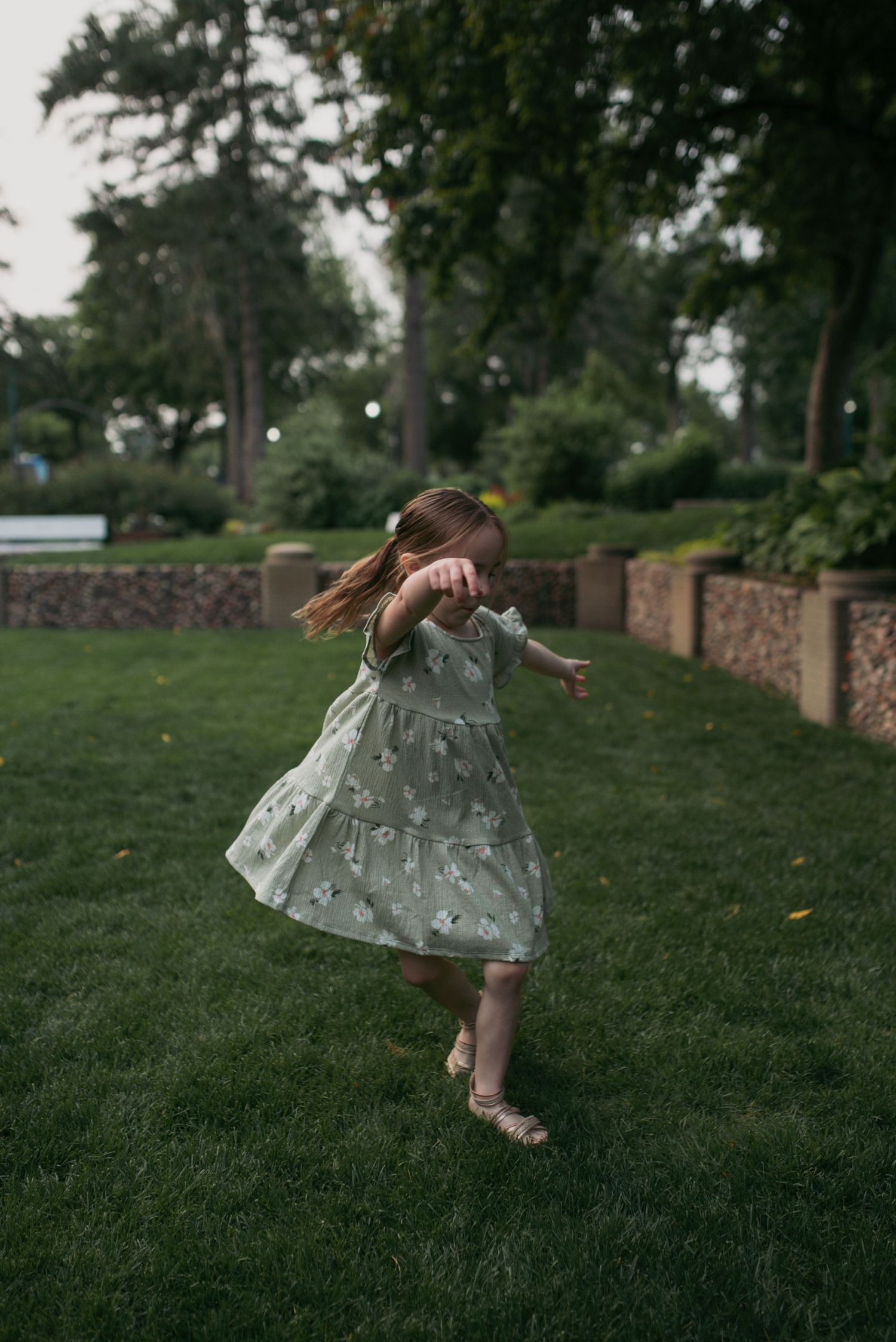 A young girl in a mint green floral dress twirls energetically on green grass in a lush park setting.