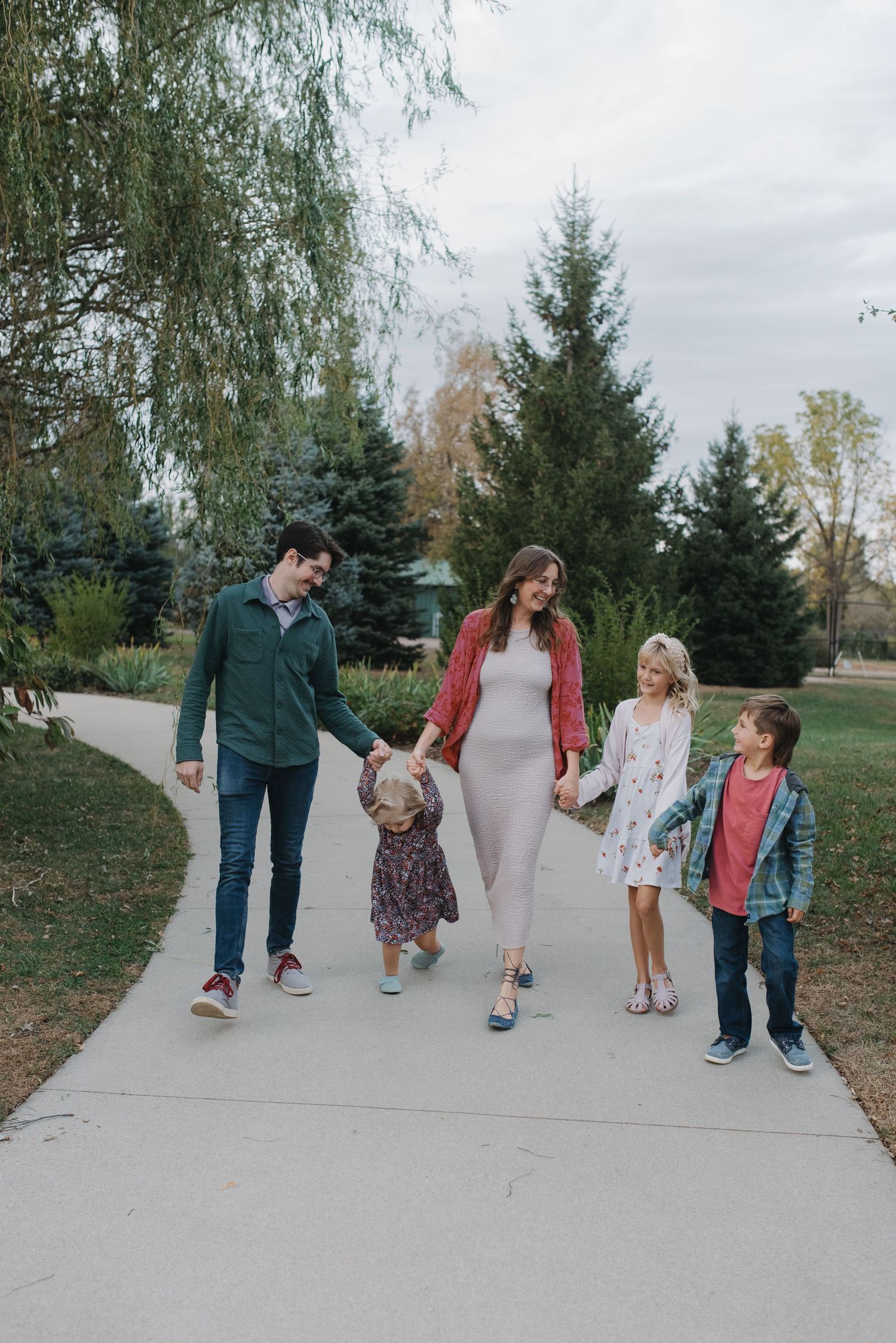 Joyful family walking together on a park path with evergreen trees in background.