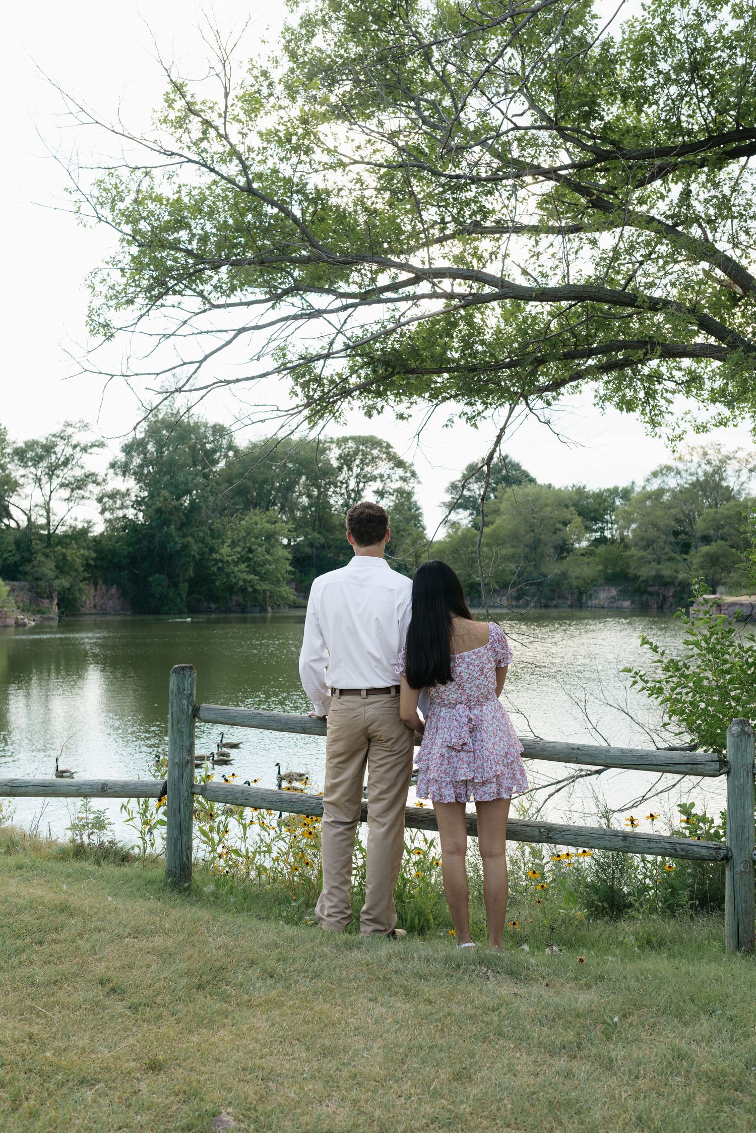 A couple stands hand-in-hand gazing at a tranquil lake with geese, framed by lush trees.