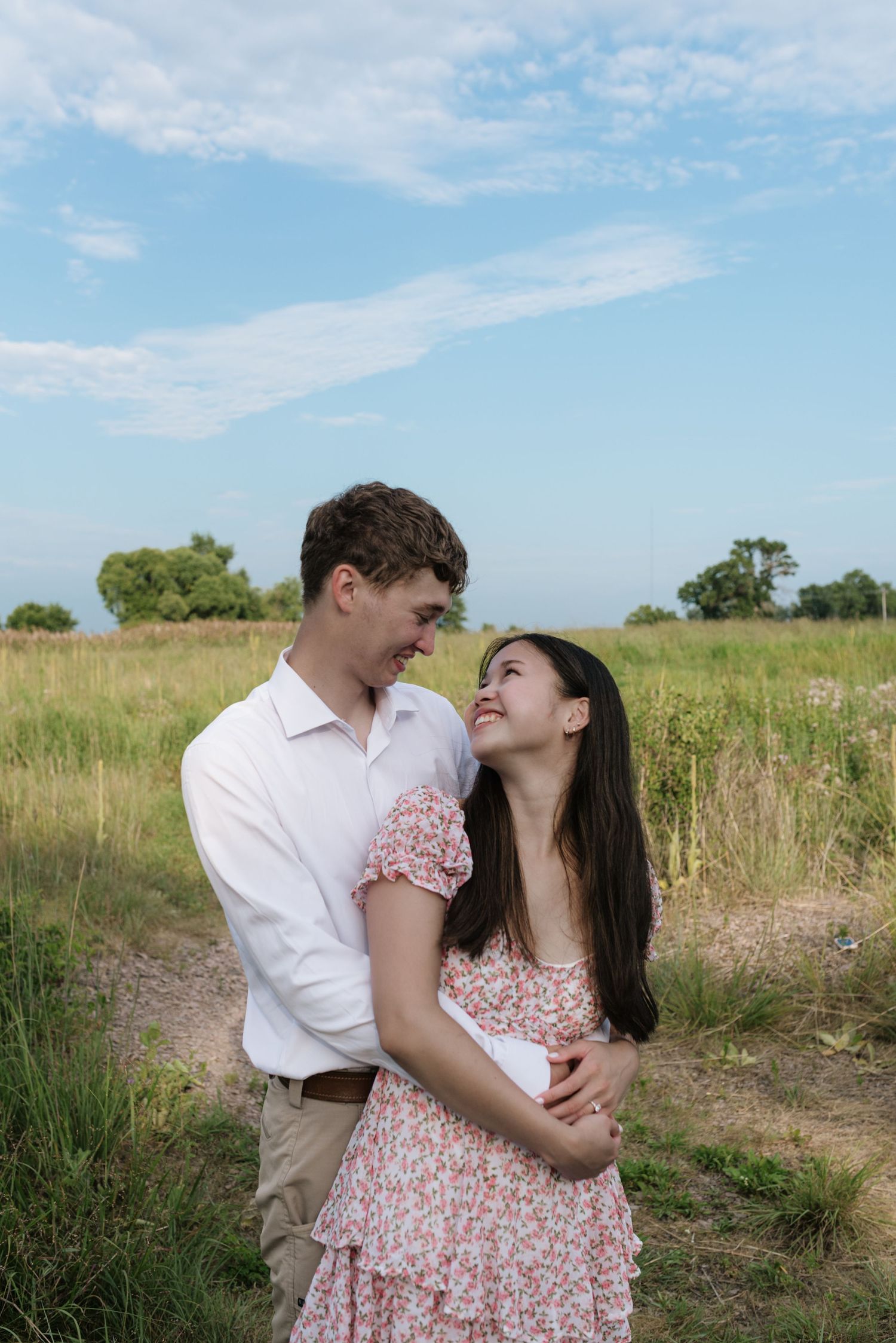 Laughing couple embracing and gazing at each other in a green meadow under a partly cloudy sky.