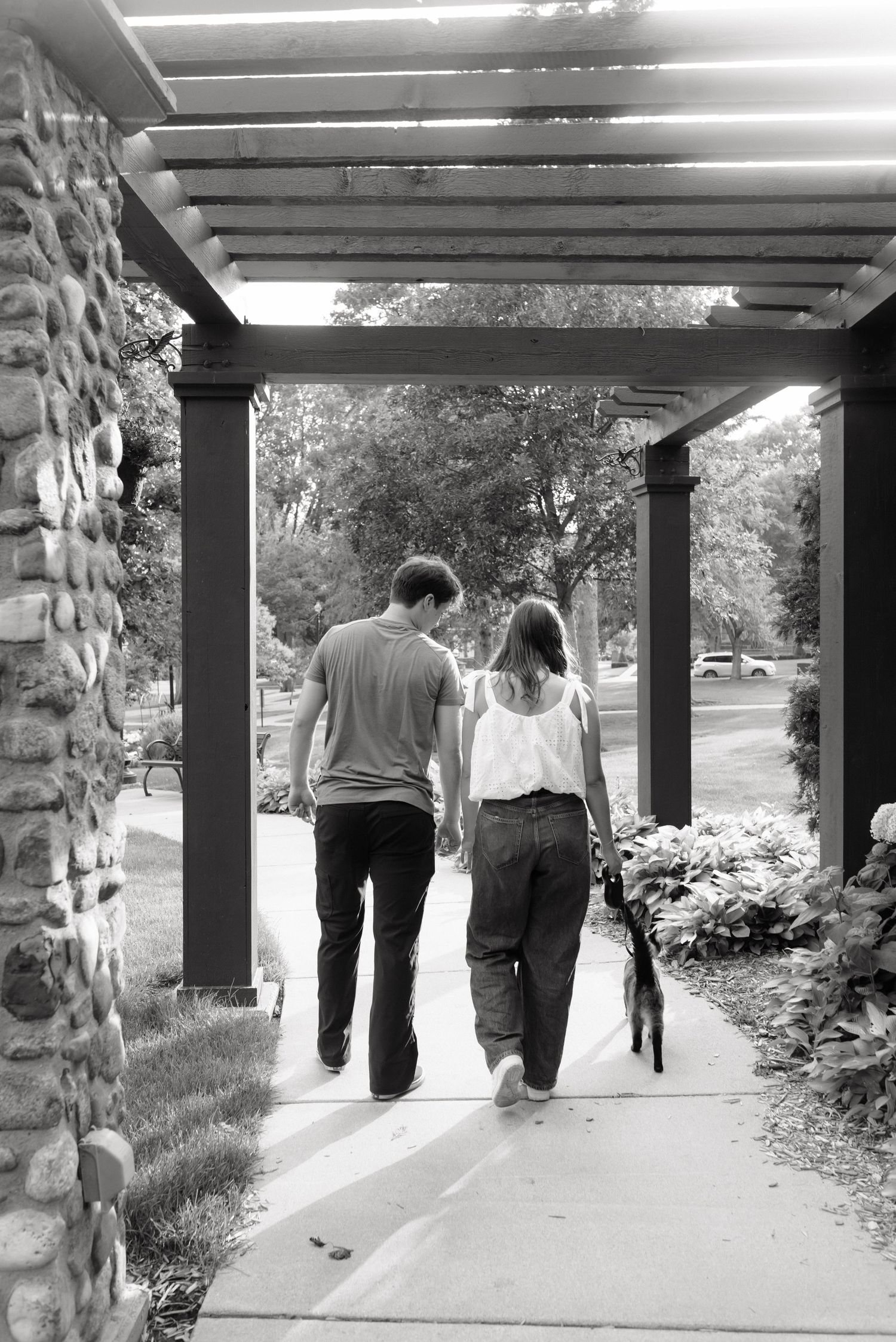 Black and white photo of a couple walking hand in hand under a pergola with a dog on a tree-lined path.